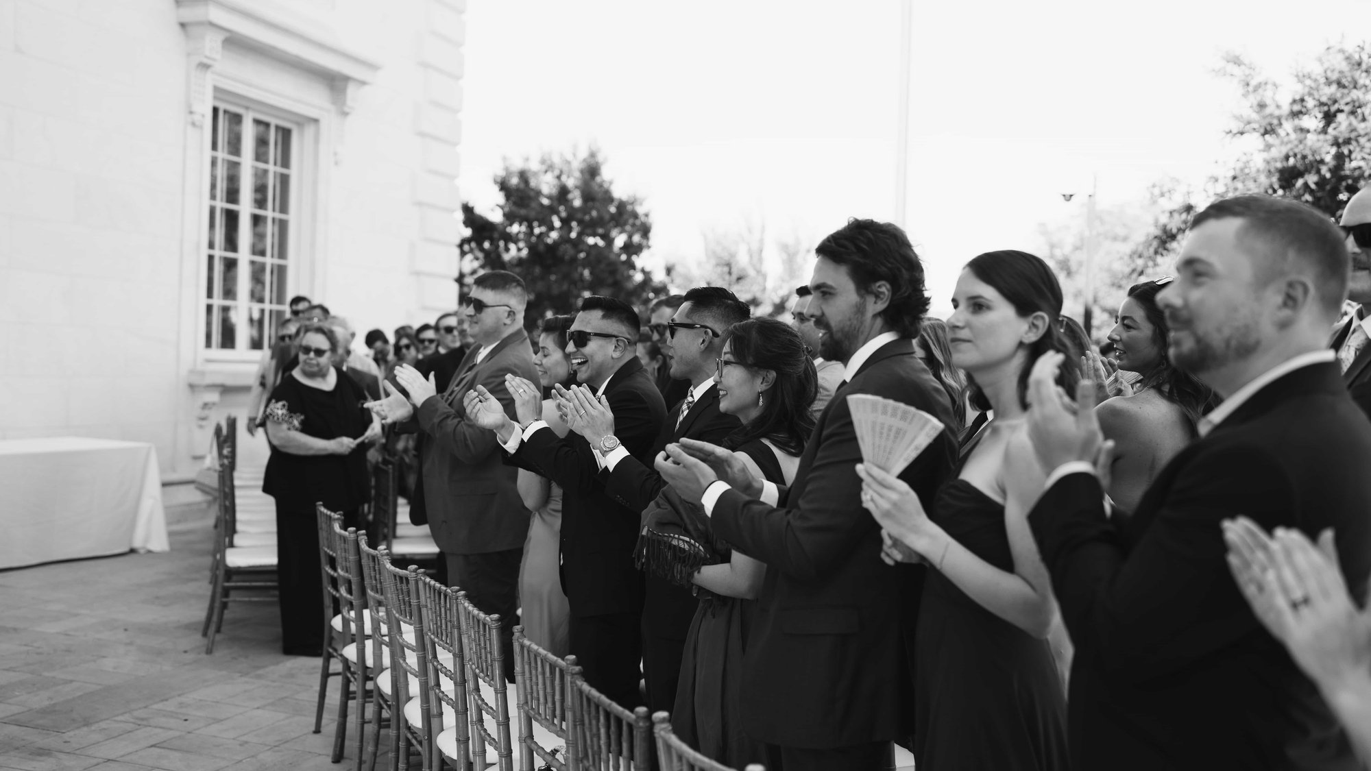 Black and white, guests on their feet applauding, an open fan raised in the middle of the row