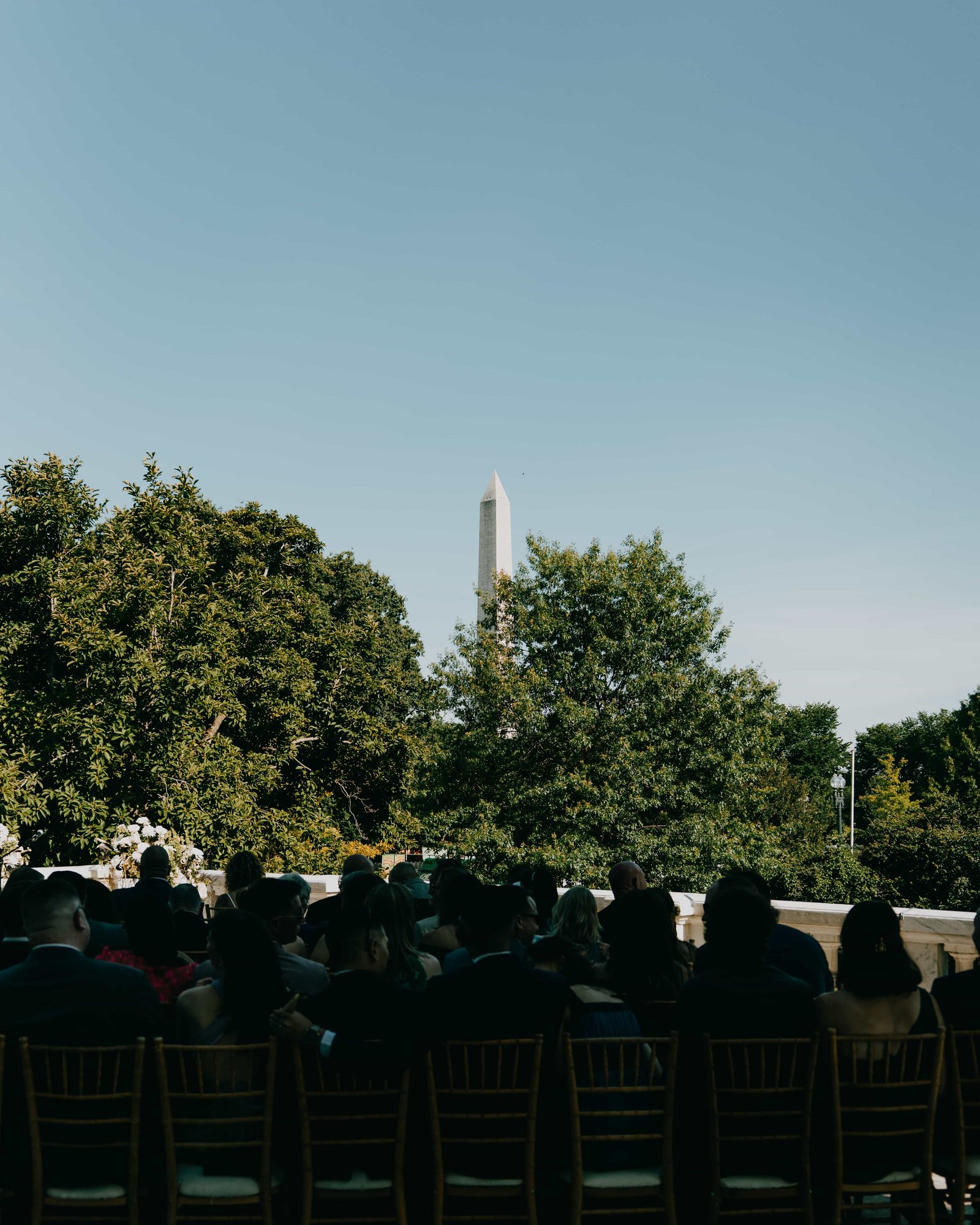 The Monument over the trees in the late sun, guests gathered in the shade below