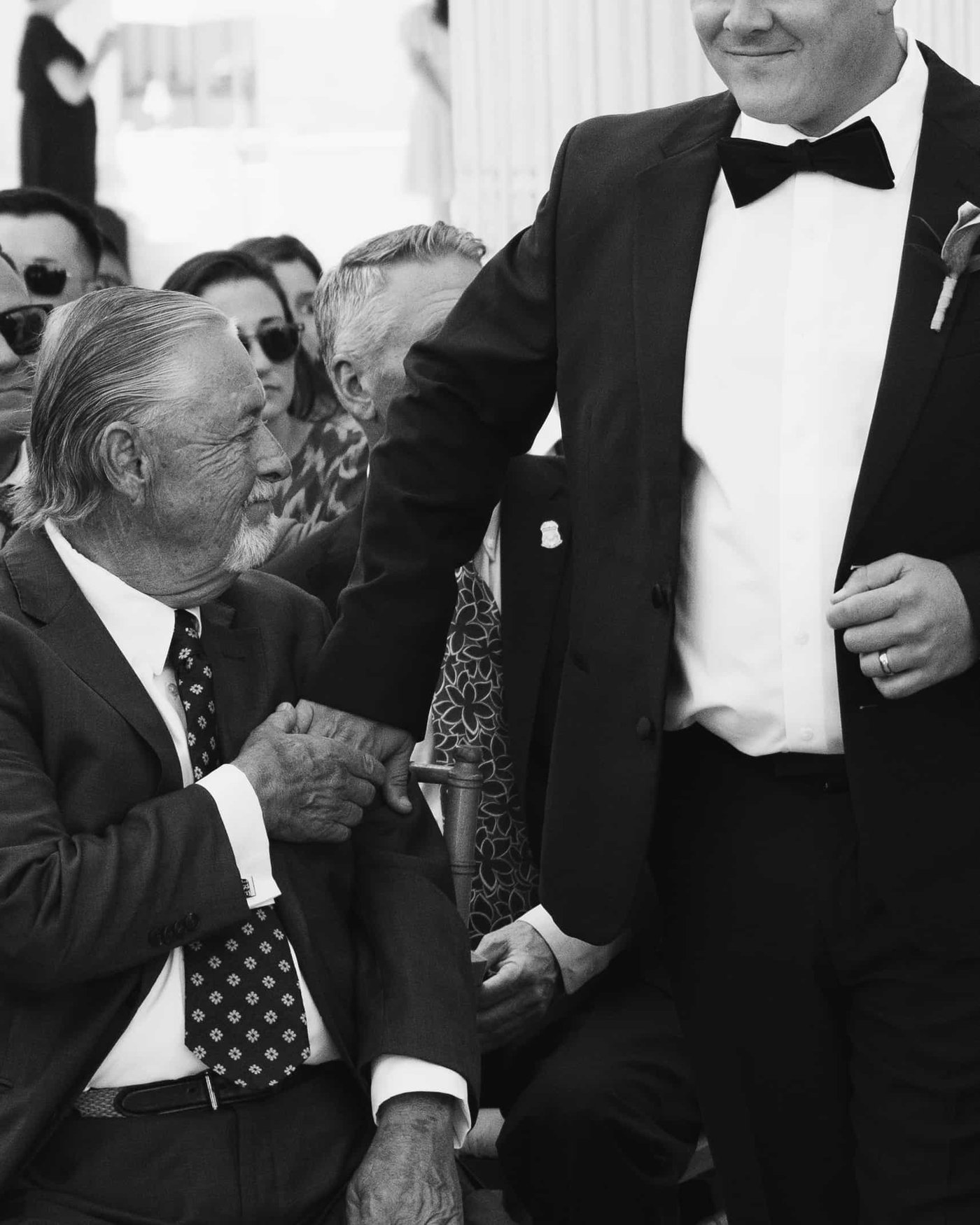 Black and white, an older guest looking up from his seat, holding fast to the groom's hand