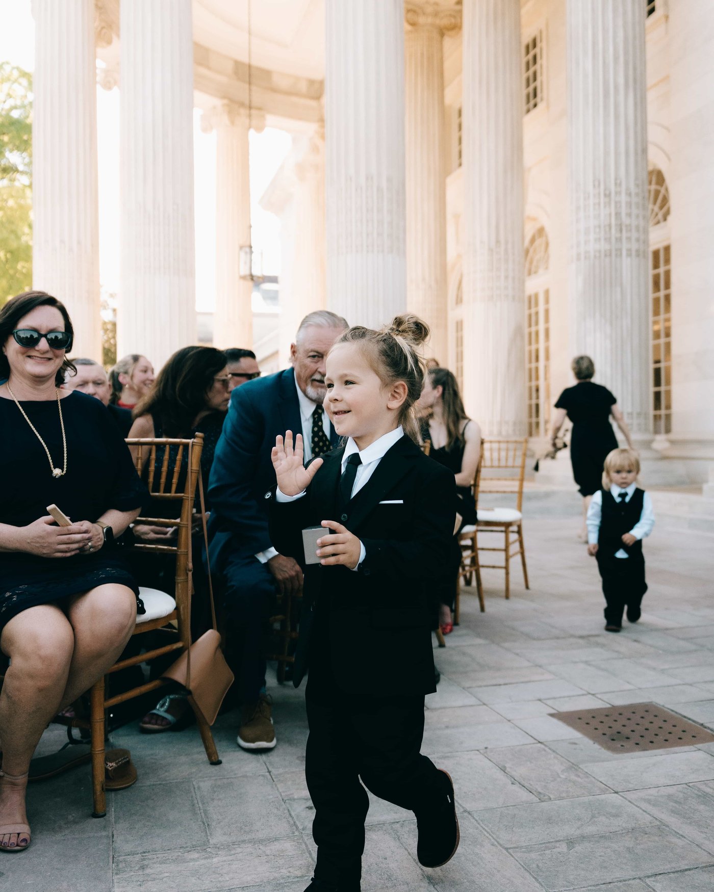 A ring bearer in a little black suit trotting down the aisle mid wave, another following far behind in his vest