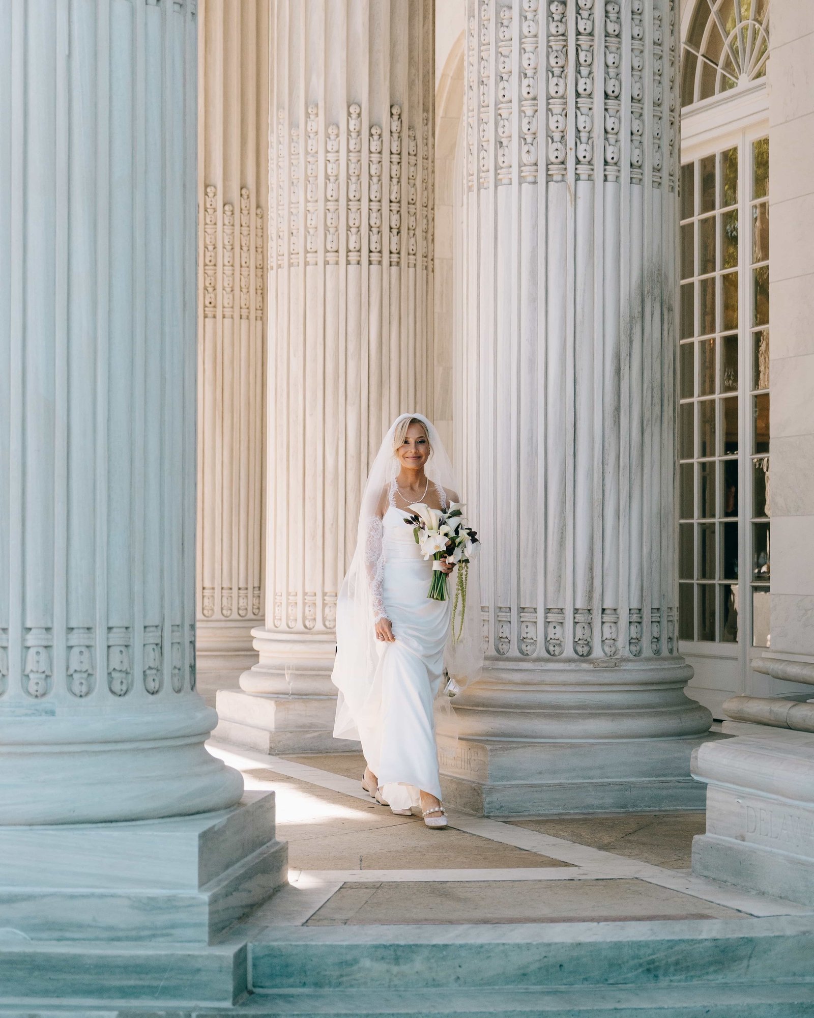 The bride carrying her callas between the towering fluted columns, her veil back and a smile already going