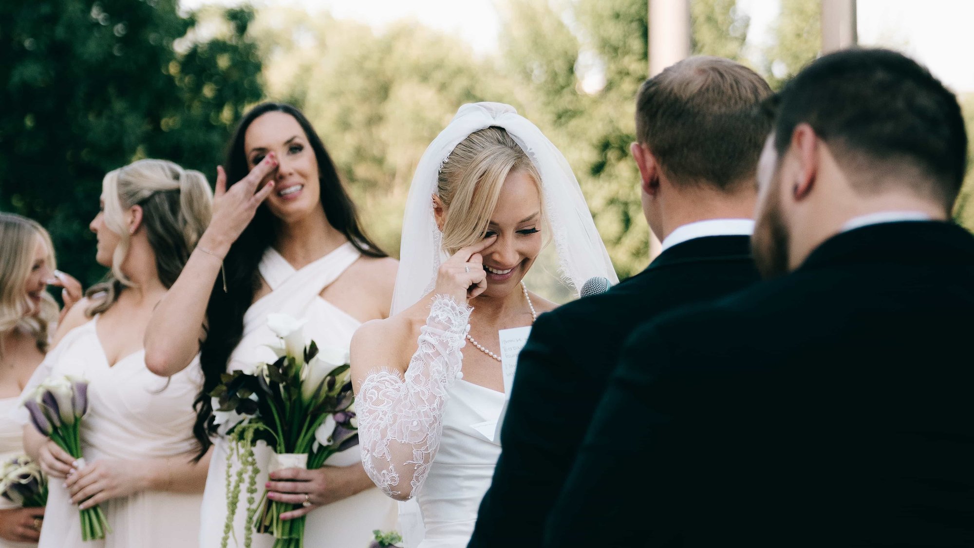 The bride laughing through a tear during the vows, a bridesmaid behind her wiping her own