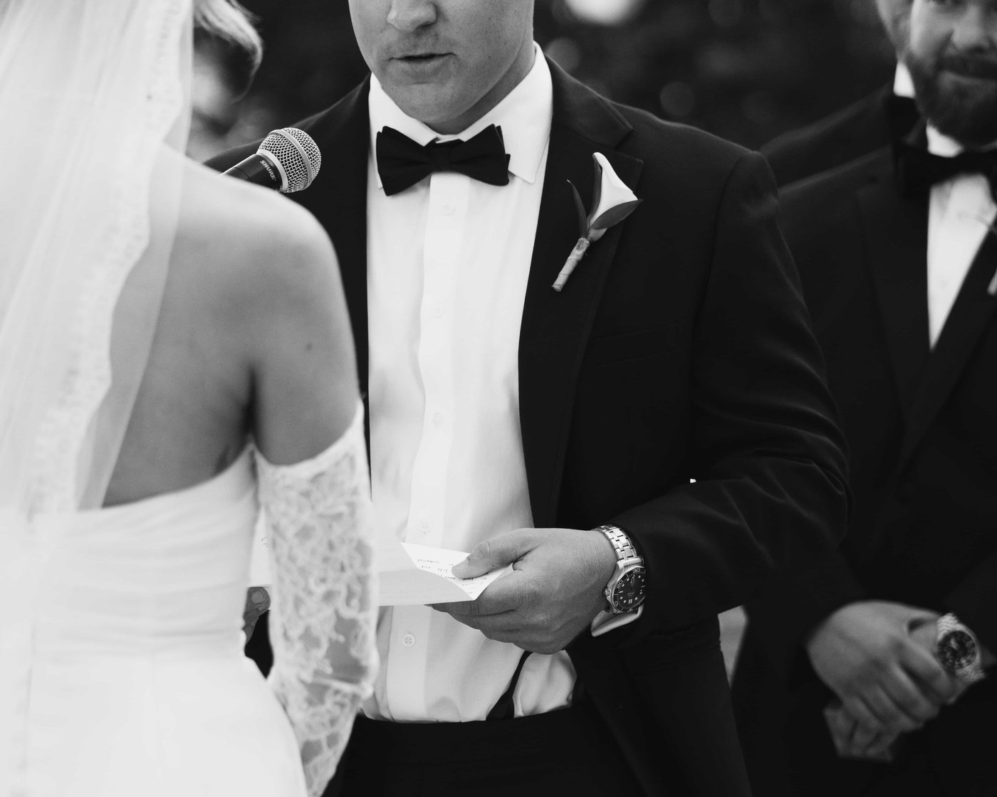 Black and white, the groom reading his vows from a folded page, a microphone held close