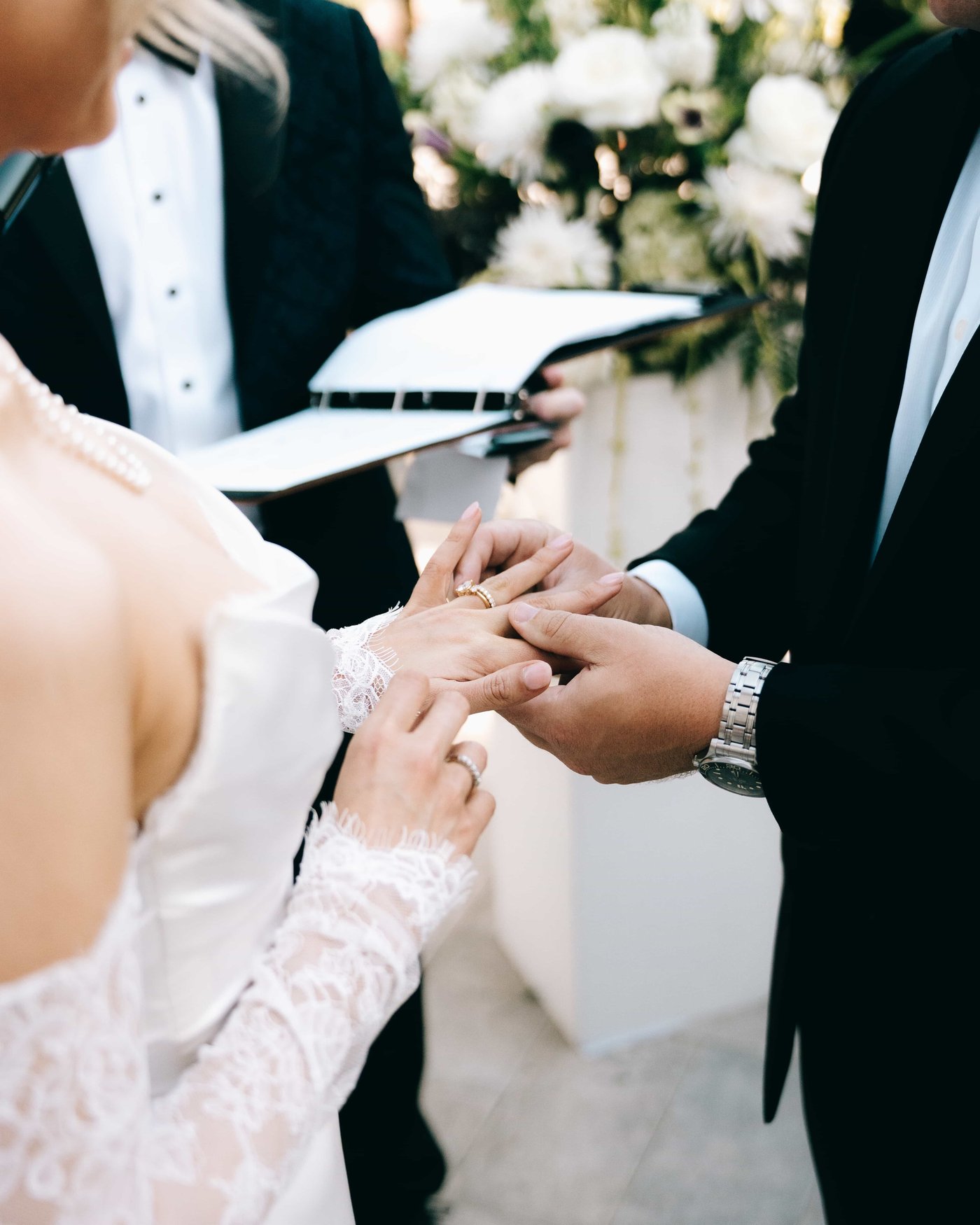 The exchange of rings, her lace sleeve against his watch, white flowers over their shoulders