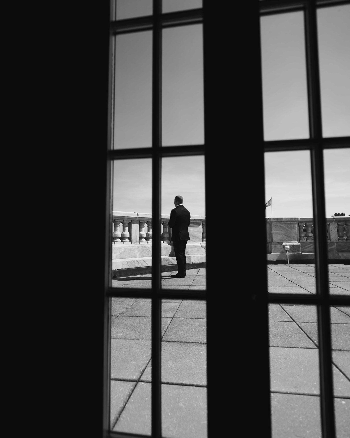 Black and white, the groom waiting alone at the balustrade, seen through the panes of the terrace doors