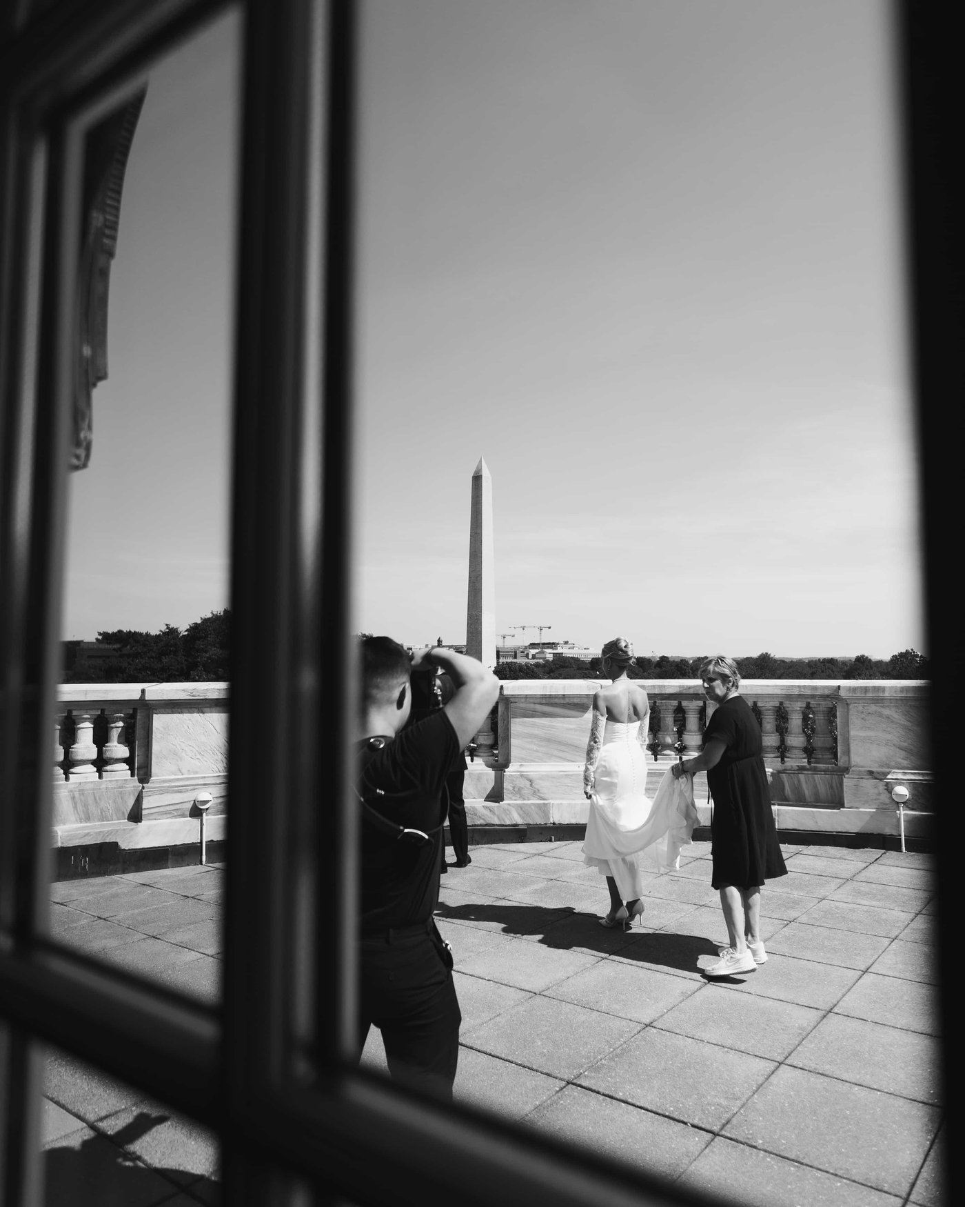 Black and white, through the glass, the bride crossing the terrace toward the Monument with her train carried behind her