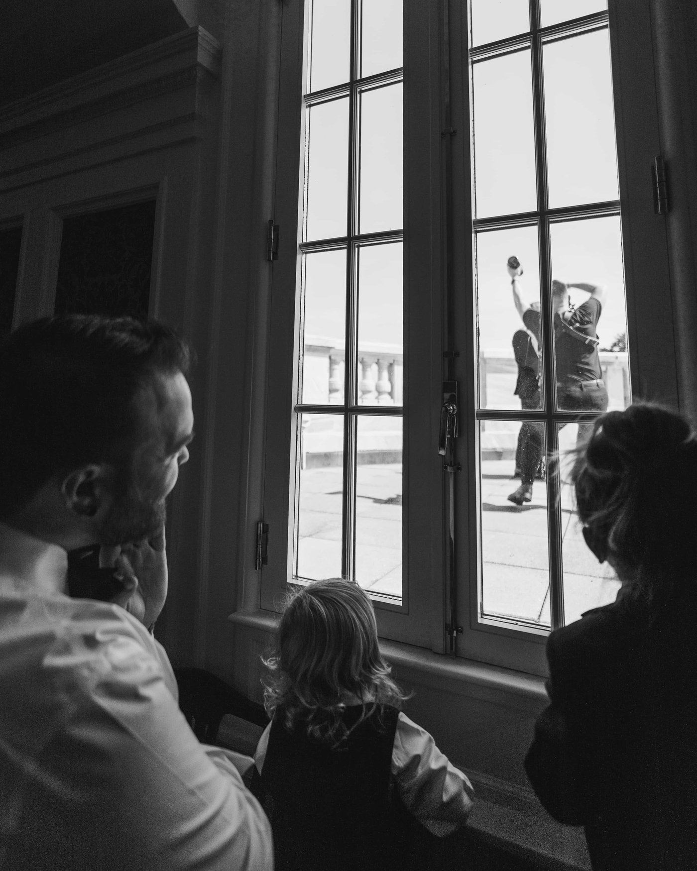 Black and white, guests pressed to the tall window, a little girl watching the celebration out on the terrace