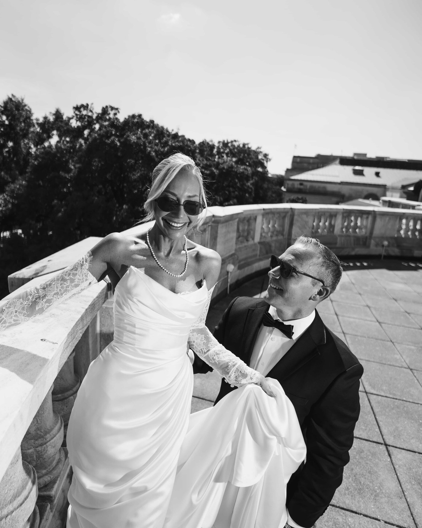 Black and white, the bride in sunglasses laughing at the balustrade while the groom looks up at her holding her train