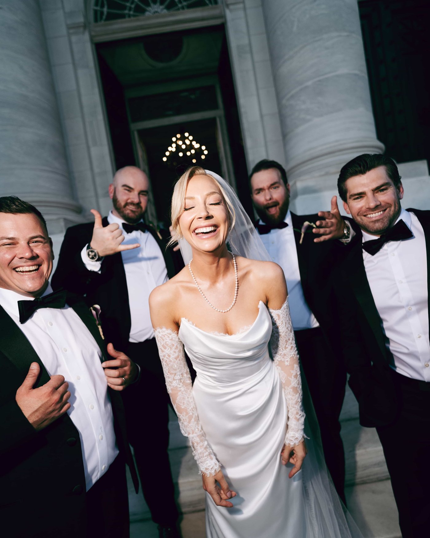 The bride laughing with her eyes closed, veil and pearls catching the flash, groomsmen mugging behind her