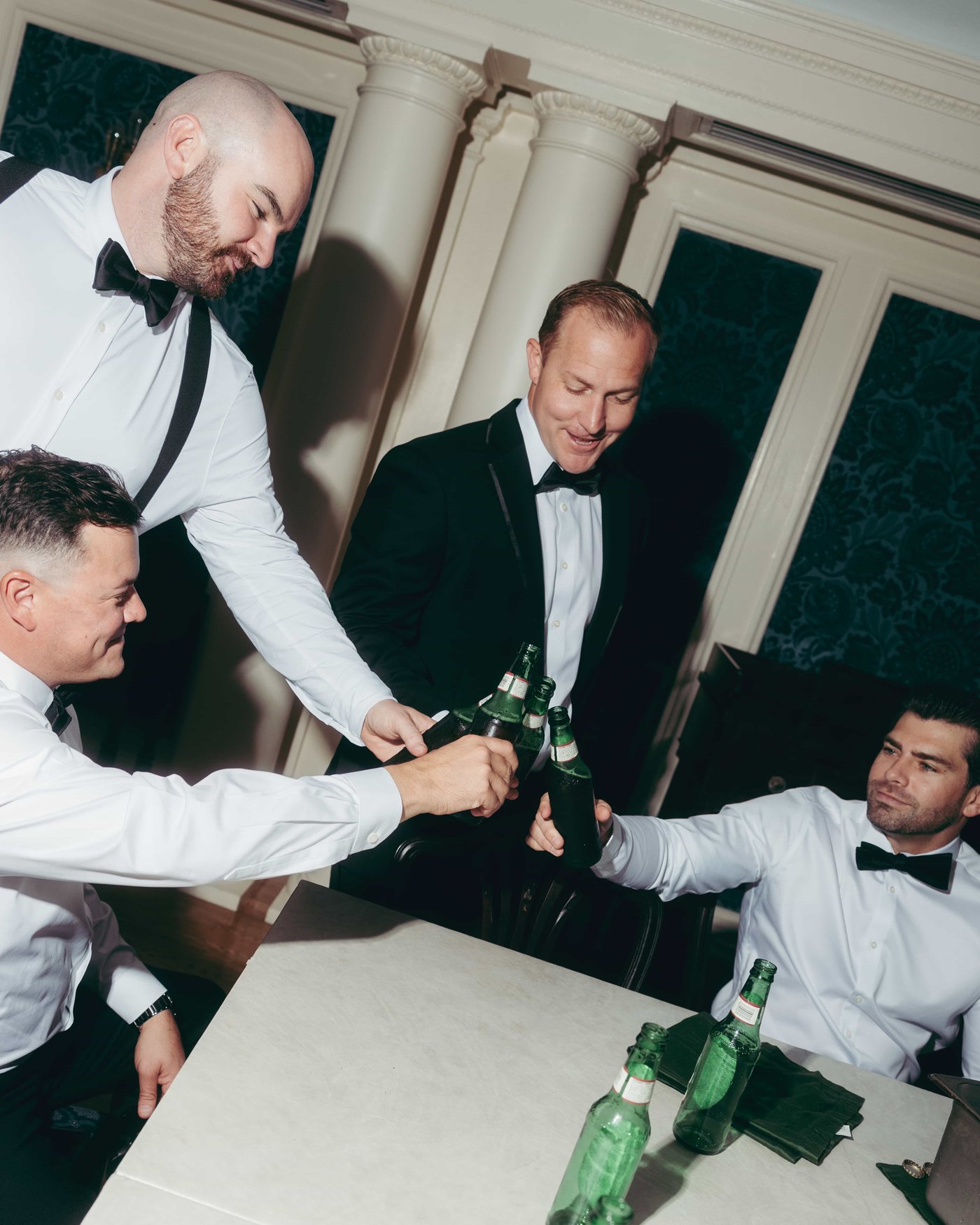 Groomsmen clinking green glass bottles across the table, one of them still in his chair