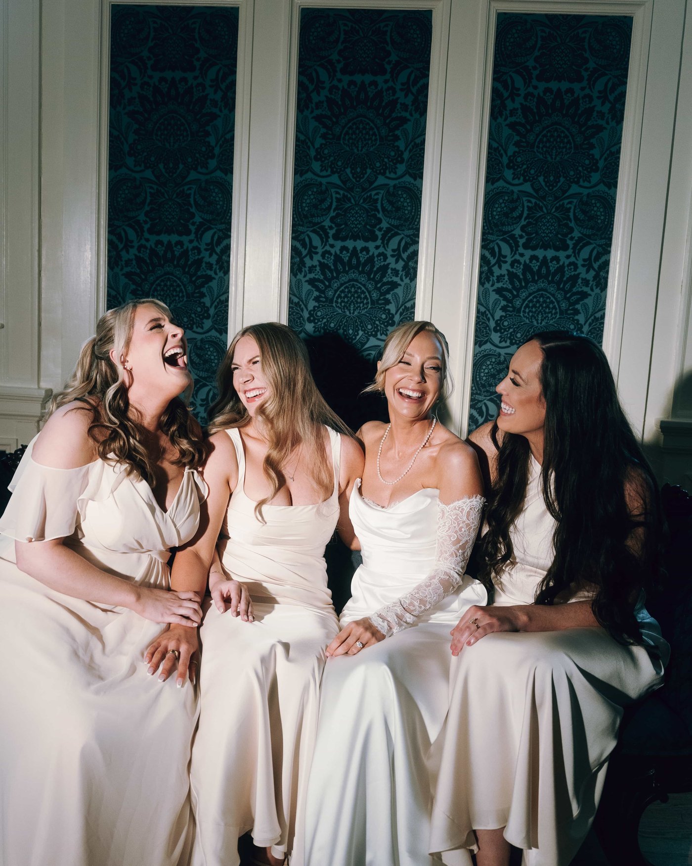The bride and her bridesmaids collapsing into laughter on a bench against the teal damask wall