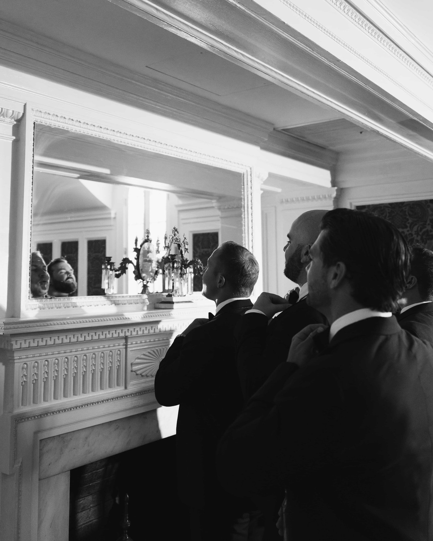 Black and white, groomsmen straightening their bow ties in the mirror above the marble mantel