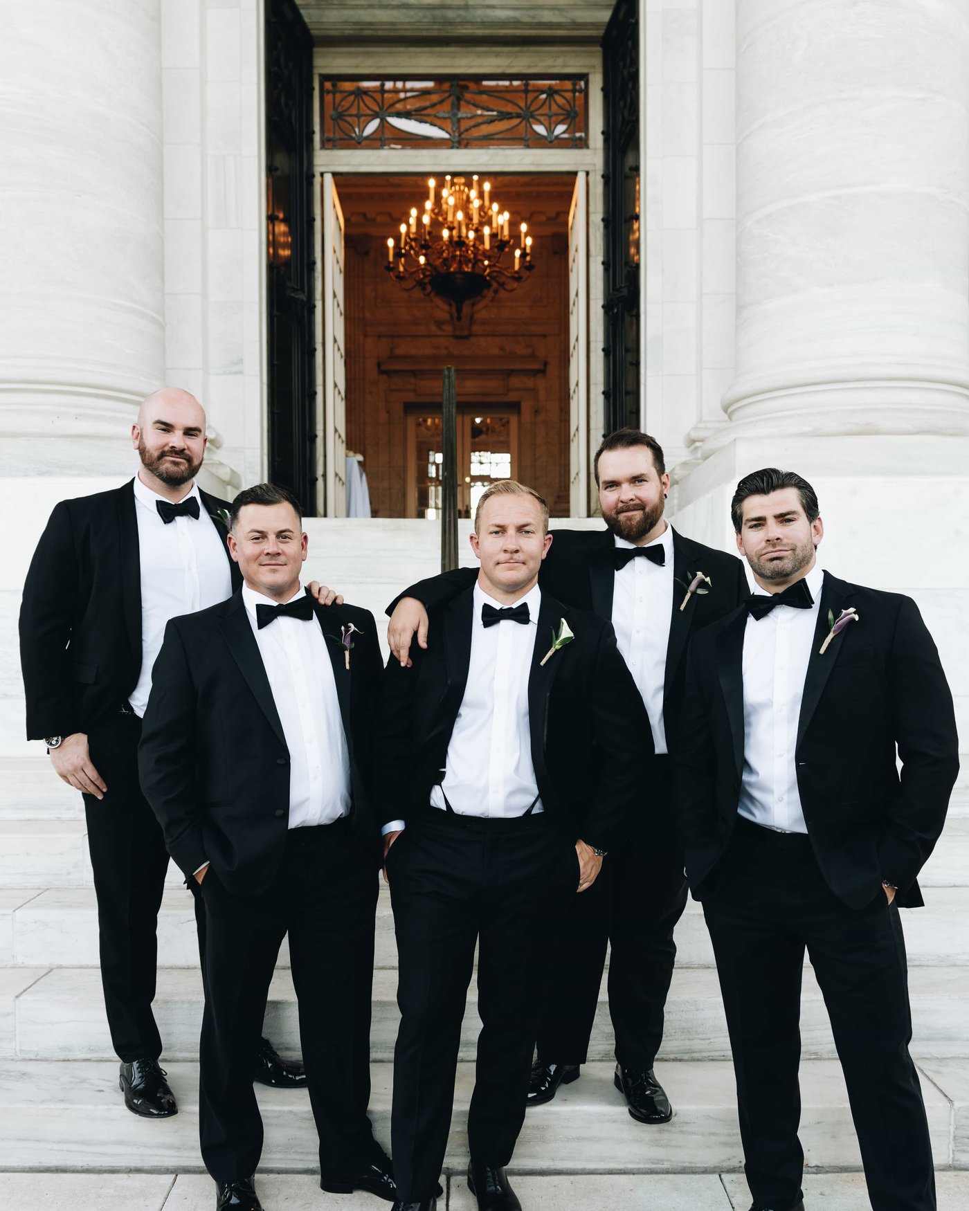 The groom and his groomsmen in black tie on the marble steps, a chandelier glowing in the doorway behind them