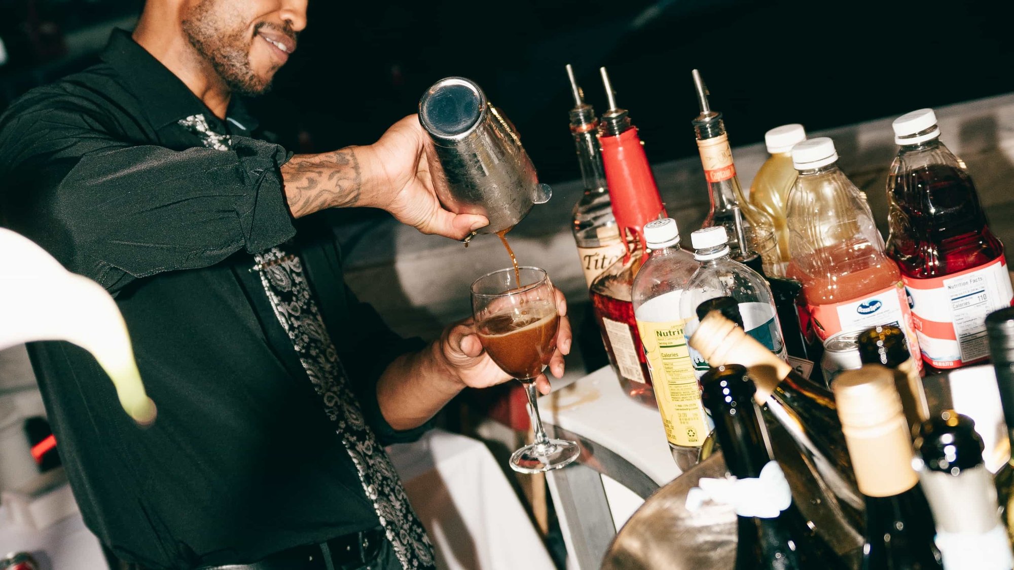 A bartender straining a cocktail into a stemmed glass, syrups and bottles crowding the rail beside him