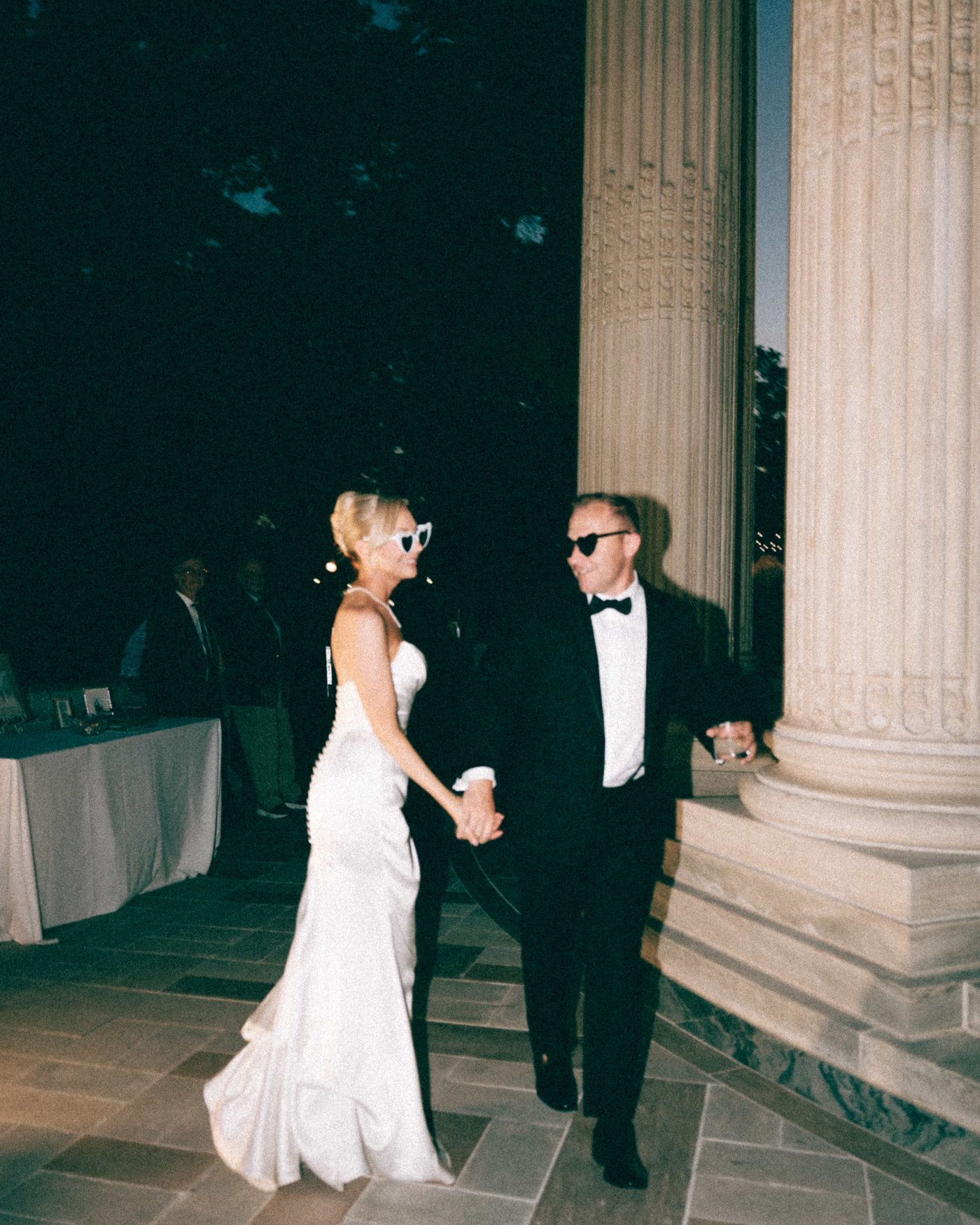 Sunglasses on after dark, the bride leading the groom past the columns by the hand