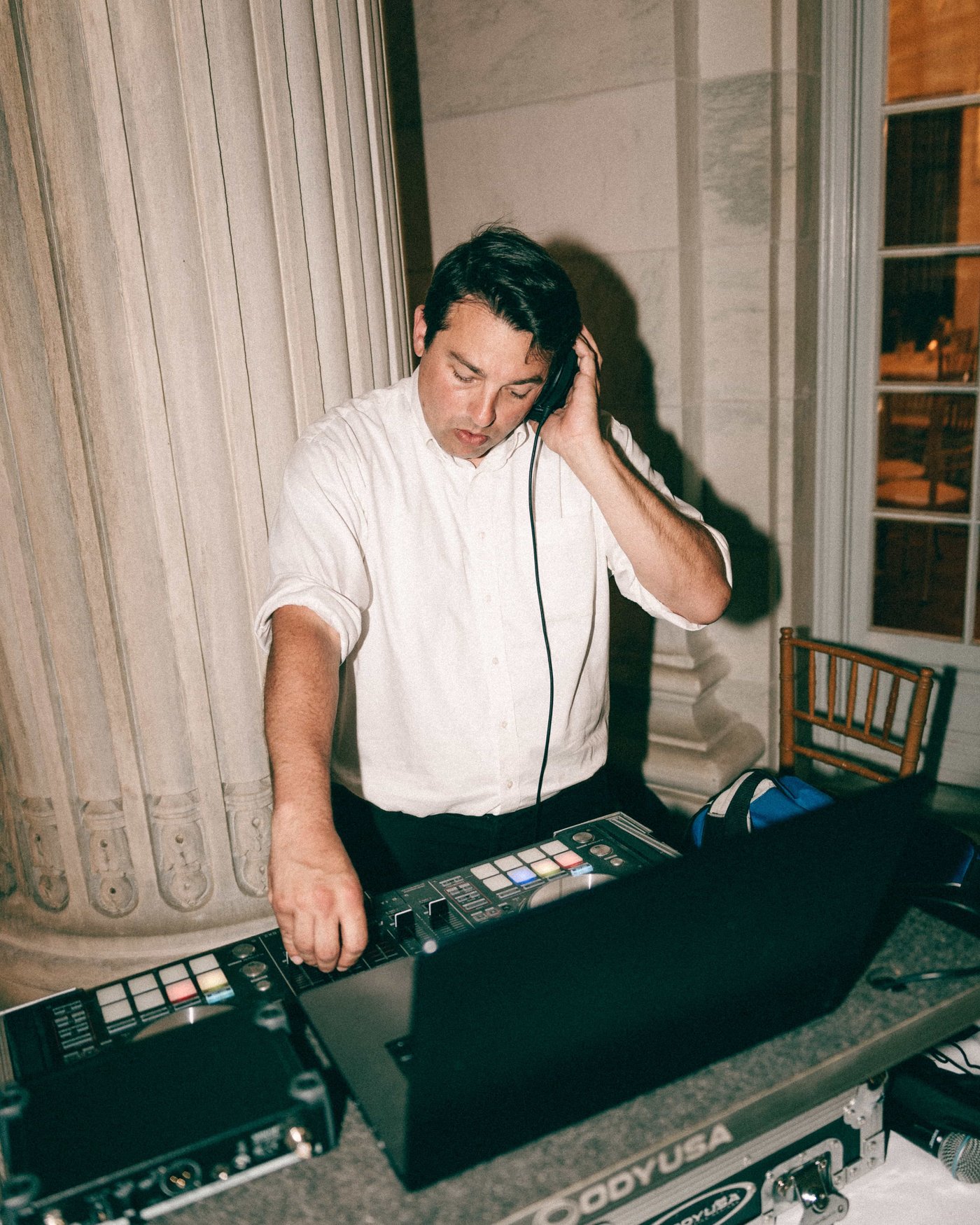 The DJ working the pads with one hand on his headphones, tucked in beside a marble column