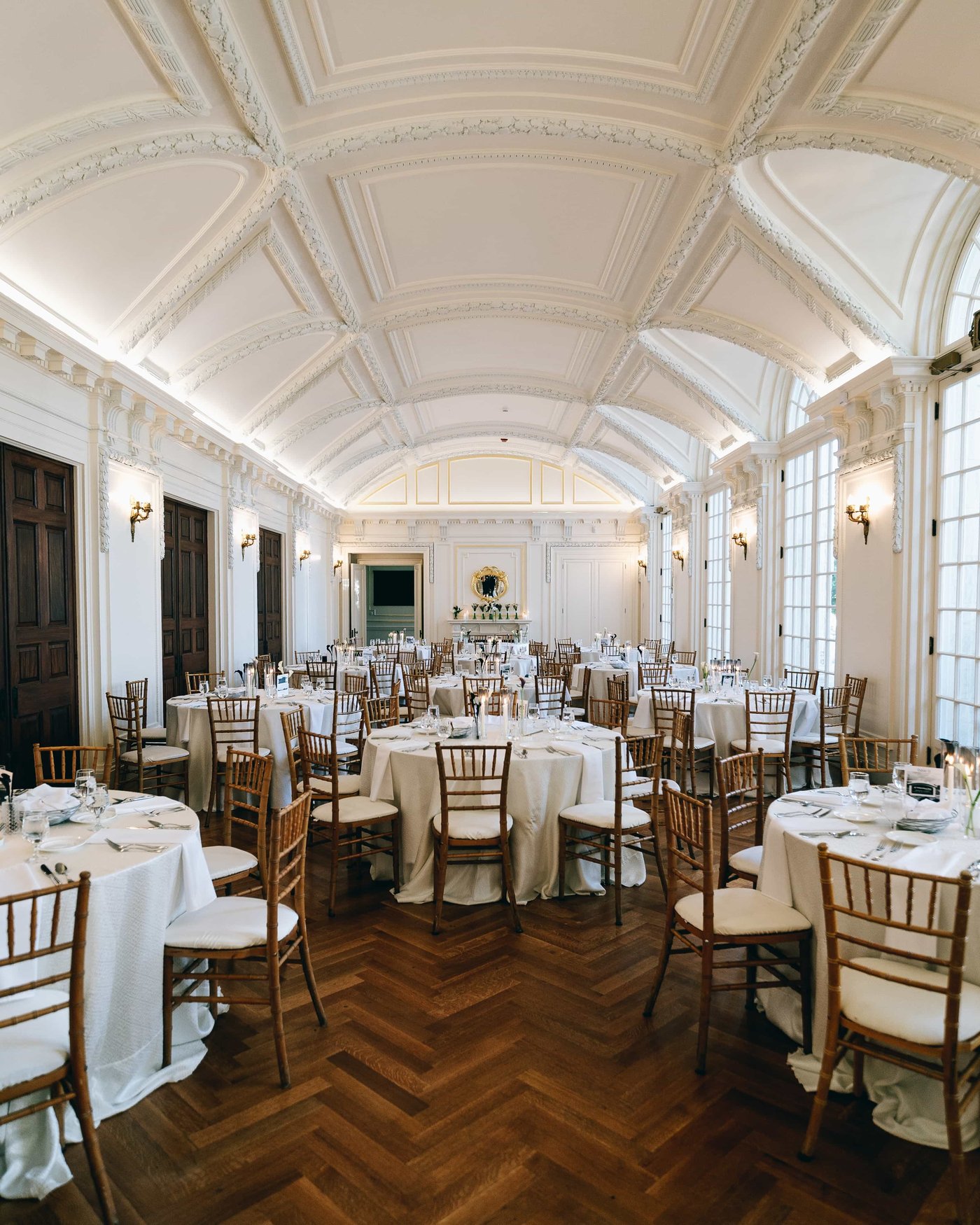 The hall set for dinner, round tables and gold chairs on the herringbone floor beneath the ornate vaulted ceiling