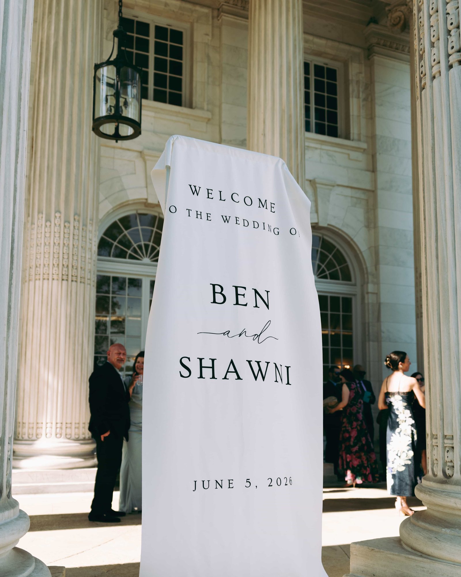 A draped fabric sign welcoming guests to the wedding of Ben and Shawni, arrivals passing between the columns behind it