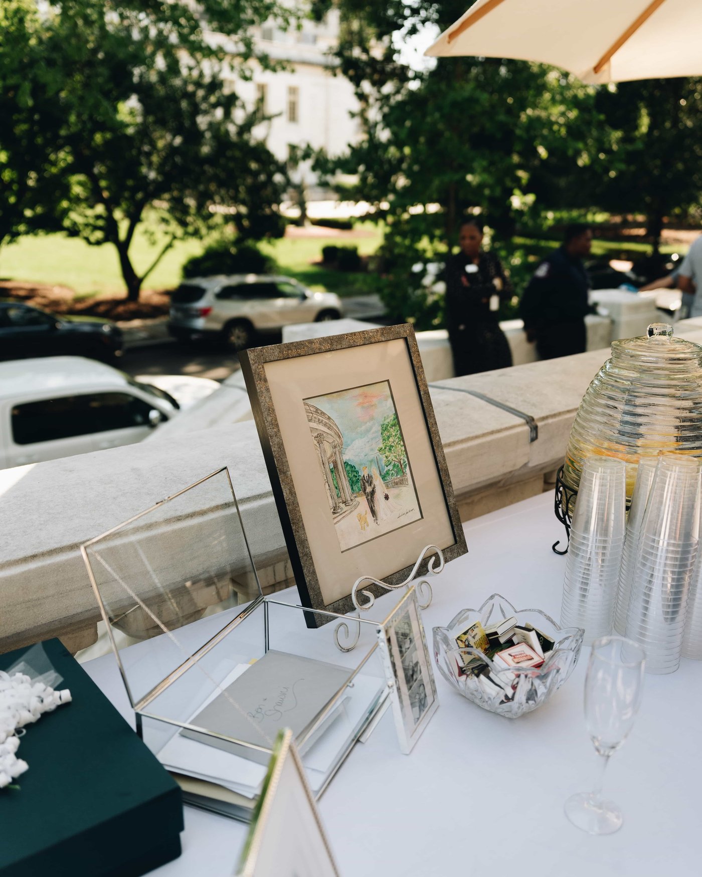 A framed watercolor propped on the gift table beside a glass card box, envelopes and a bowl of favors