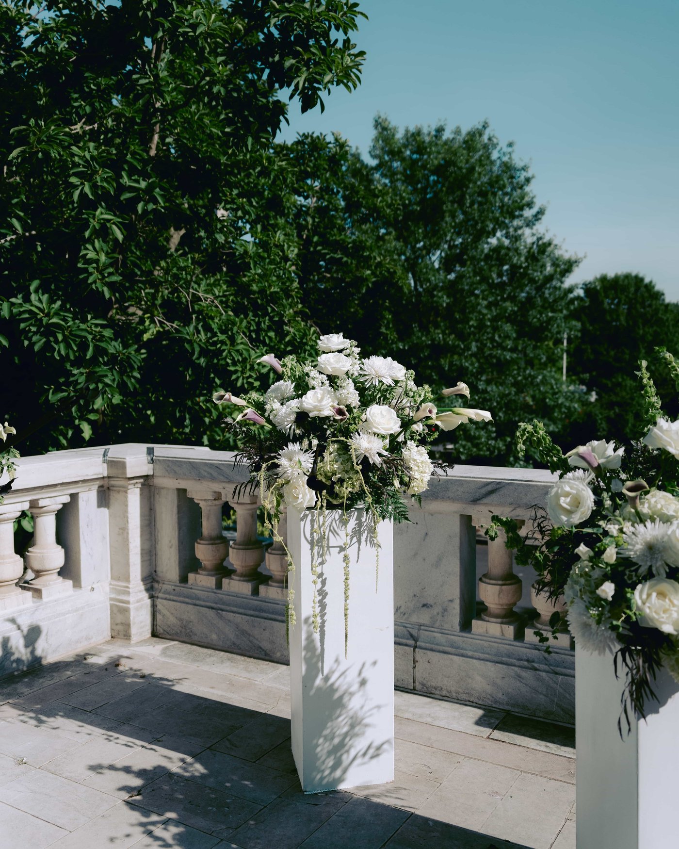 Arrangements of white roses, callas and chrysanthemums on tall white pedestals along the marble balustrade