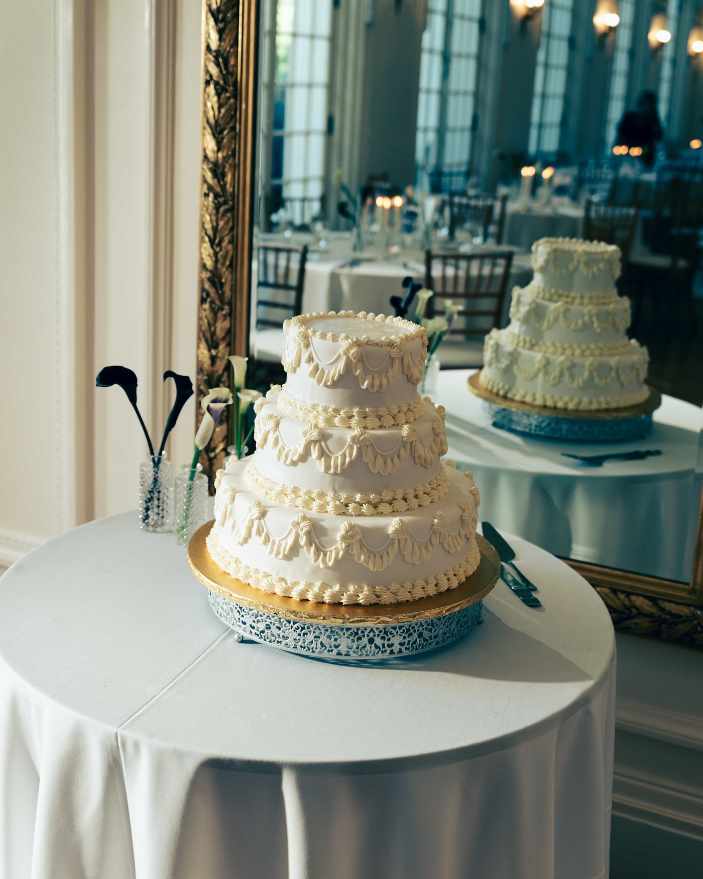 Three tiers of piped buttercream on a lace stand, doubled in the gold framed mirror behind the cake table