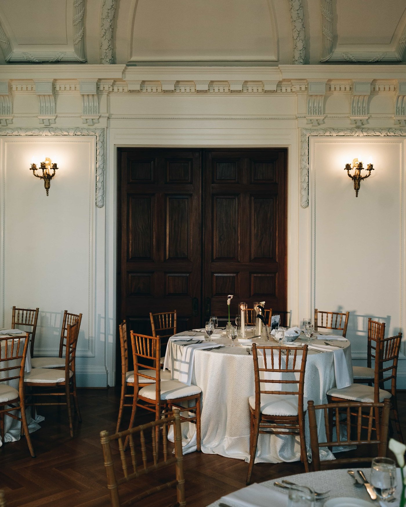 A round table set in white before the dark double doors, brass sconces lit on either side