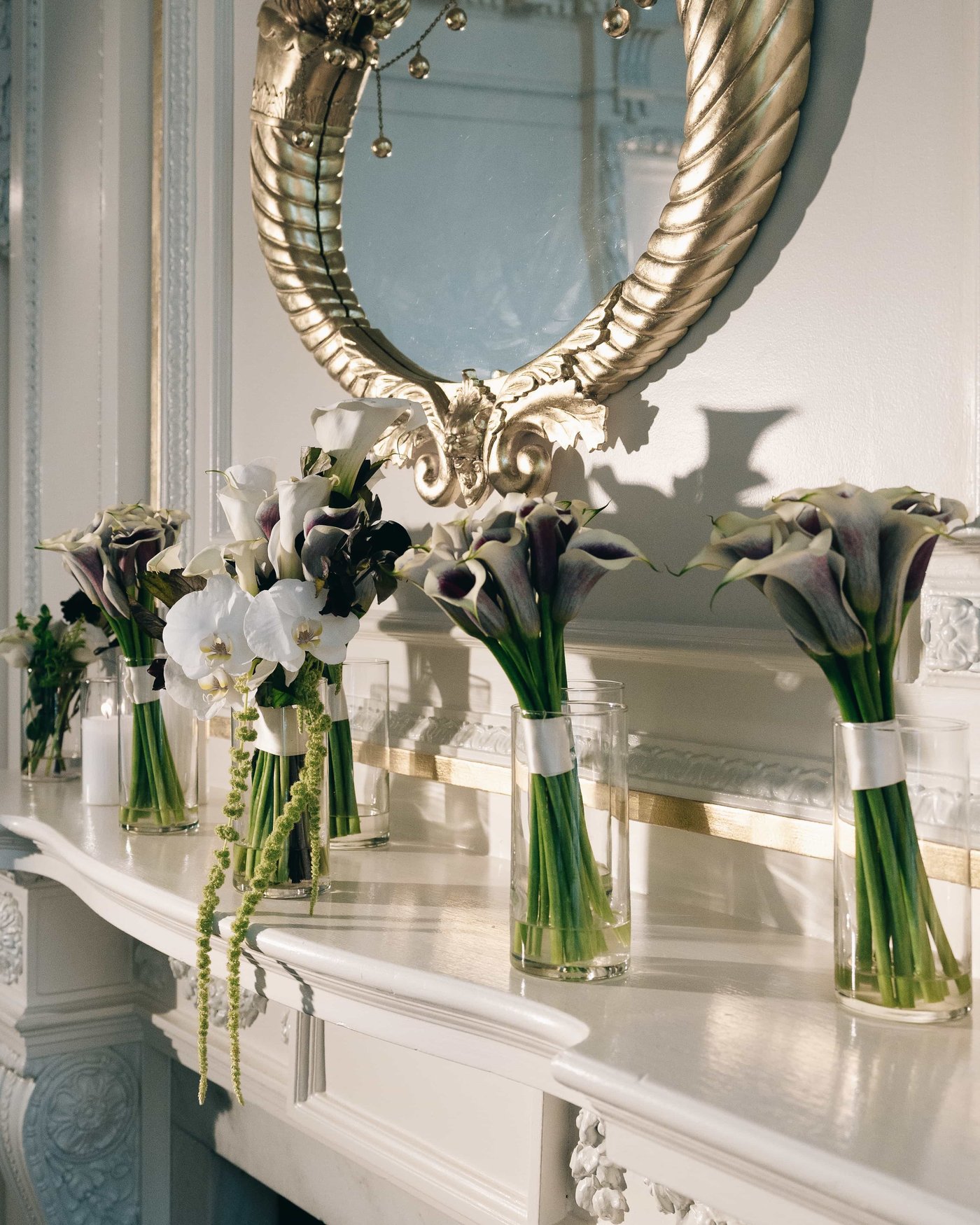 The bouquets resting in glass cylinders along a white mantel, calla lilies and orchids beneath a gilded mirror