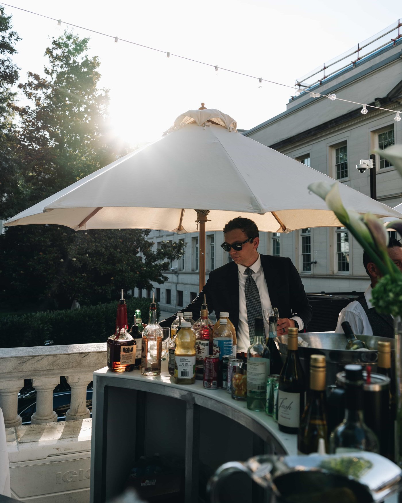 A bartender in sunglasses working under a white umbrella, bottles crowded in front of him and the sun flaring behind