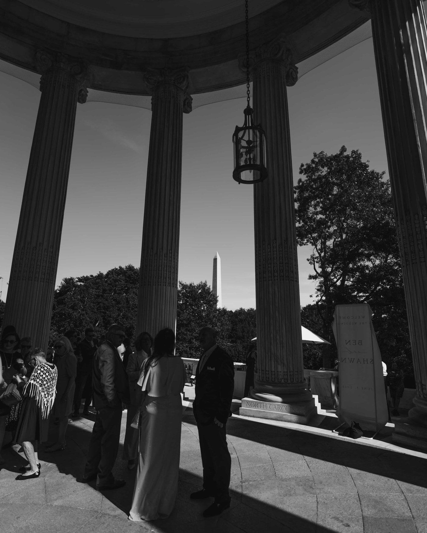 Black and white, guests gathered beneath the towering columns of the portico, a lantern overhead and the Monument in the distance