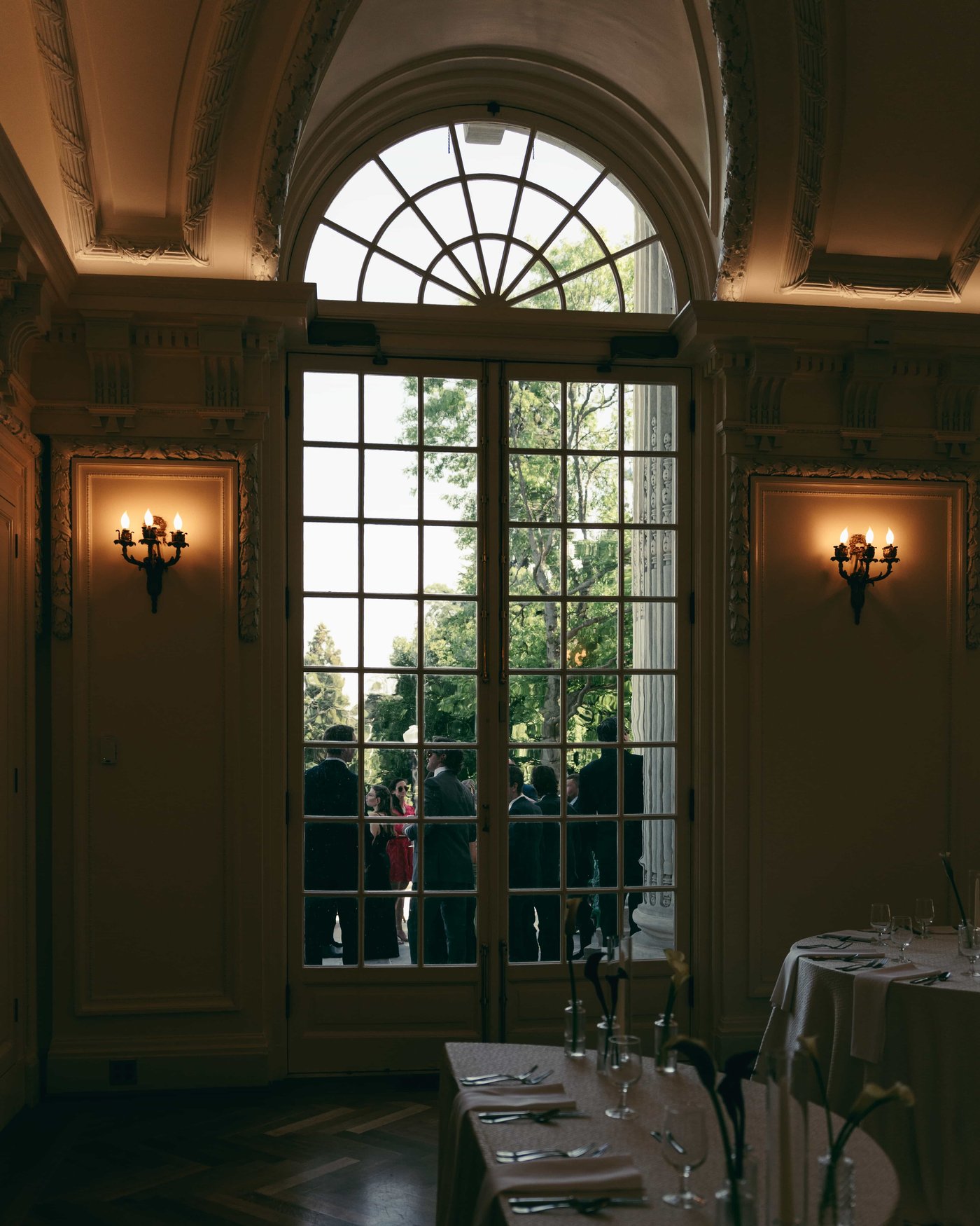 Tables set beneath a tall fanlight window, sconces lit, guests out on the sunlit terrace beyond the glass