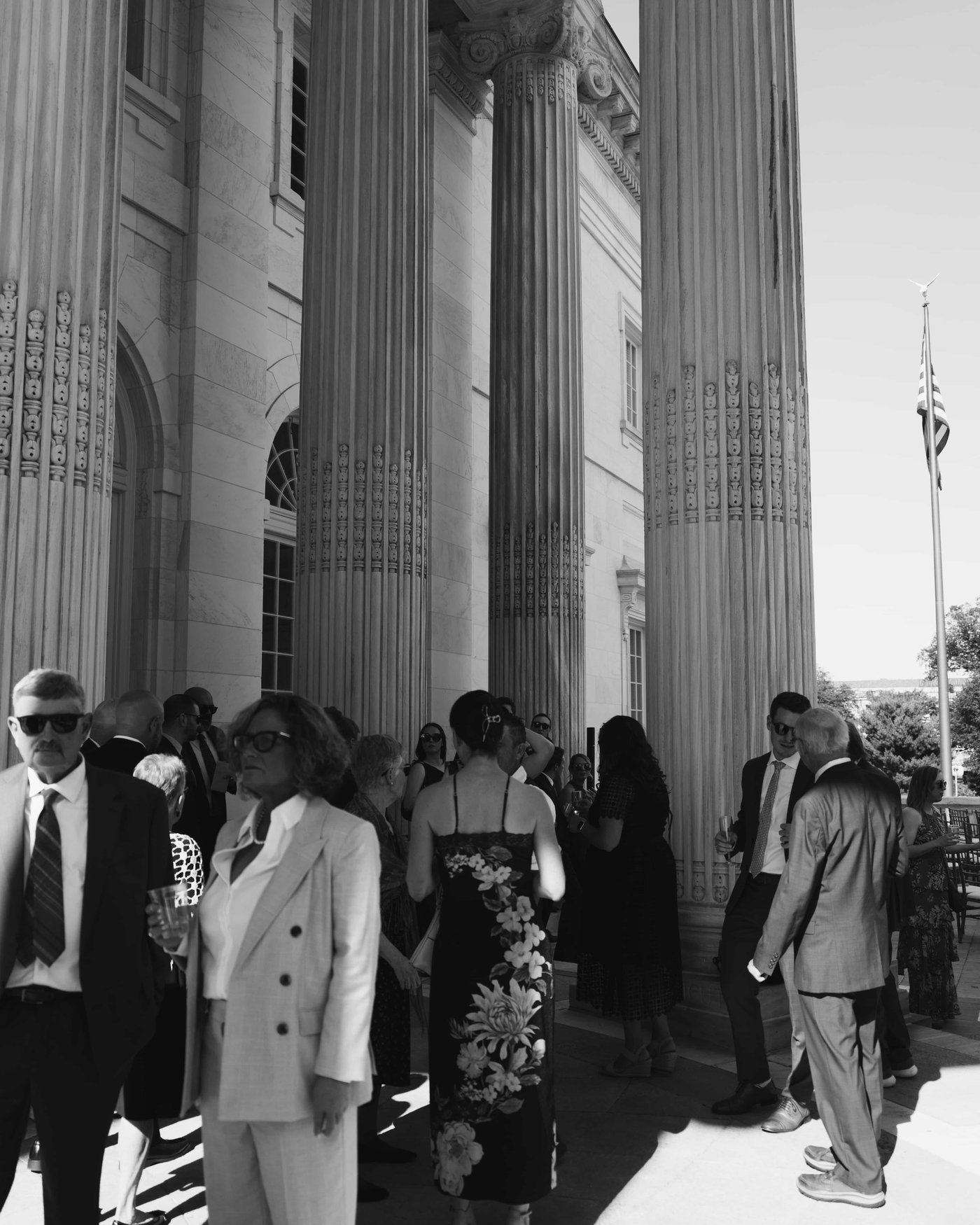 Black and white, guests in cocktail attire mingling among the giant fluted columns, an American flag in the sun