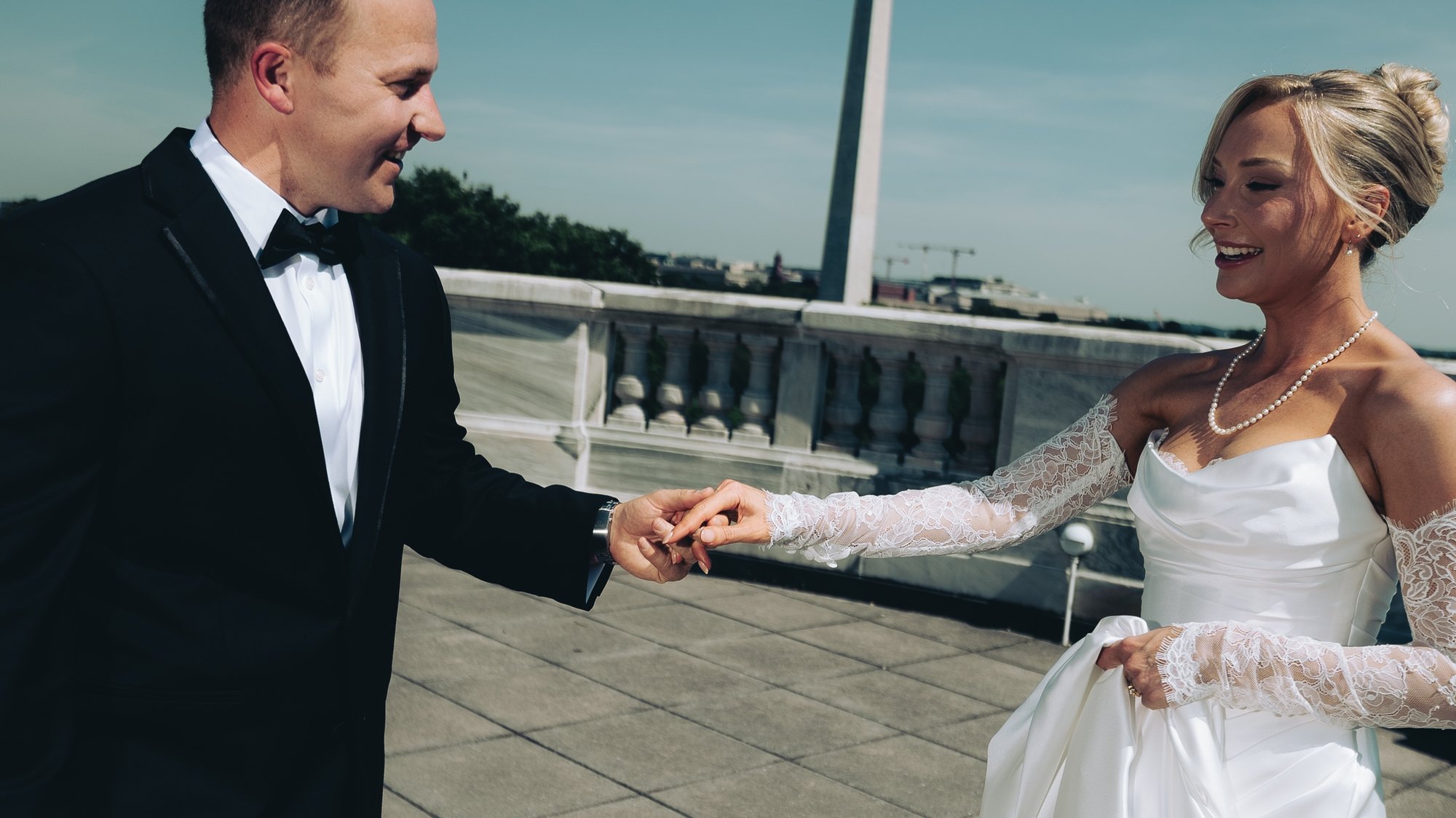 A groom leading his bride by the hand along a stone balustrade, her long lace gloves and her laugh caught mid step