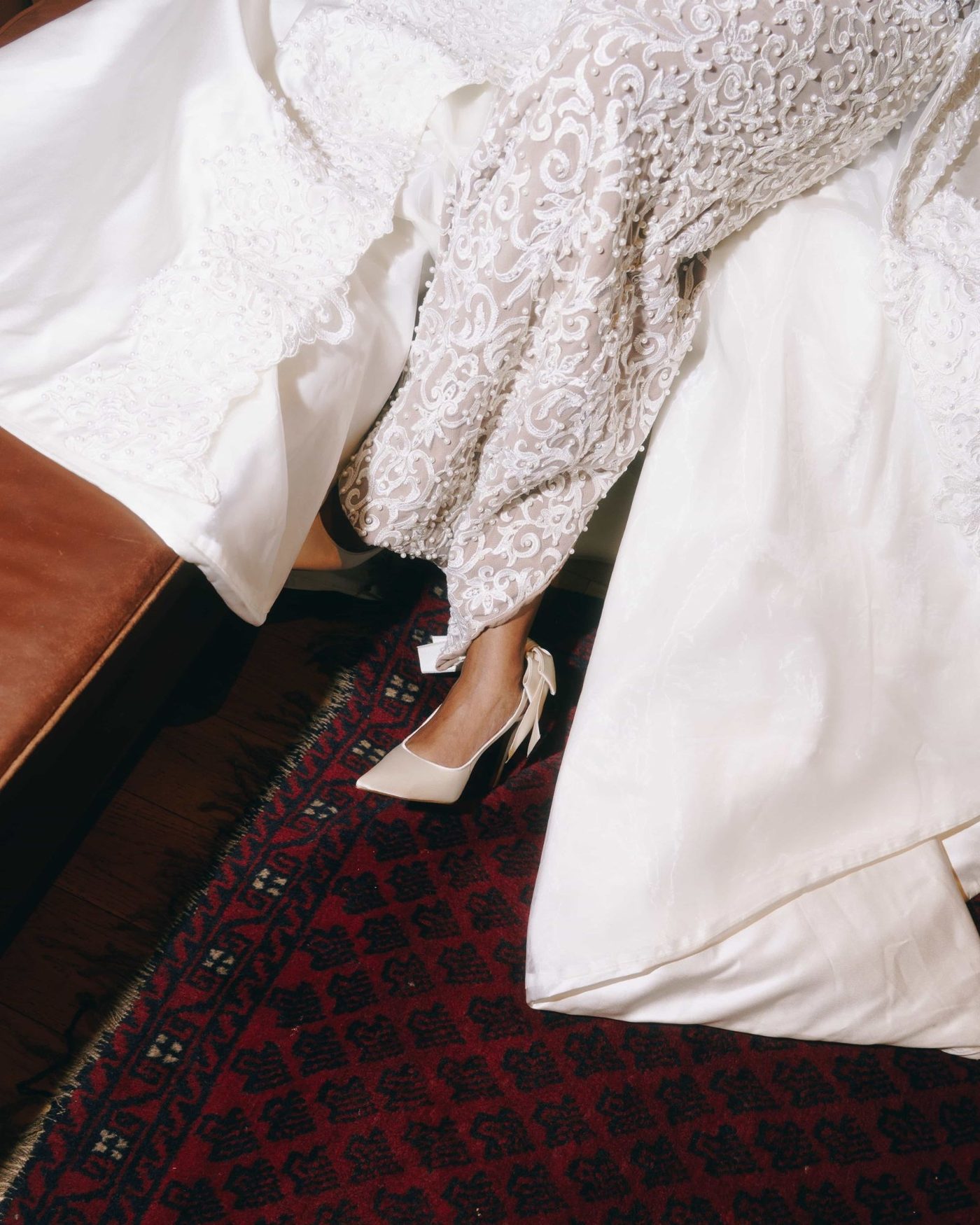 Looking down at a seated bride, the beaded lace sleeve and skirt of her gown spilling across a deep red patterned rug beside one satin heel