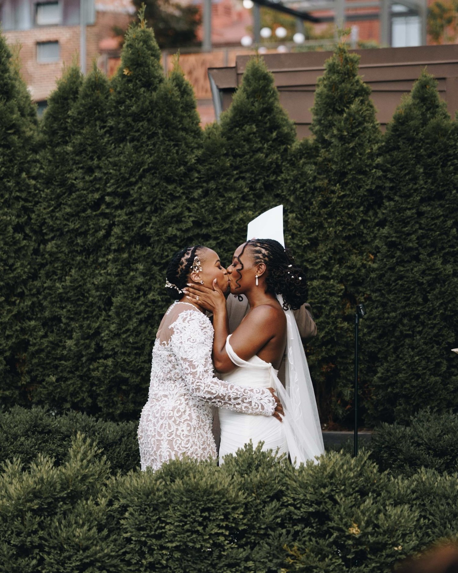Two brides kissing in front of a wall of evergreens, one veil lifting behind them