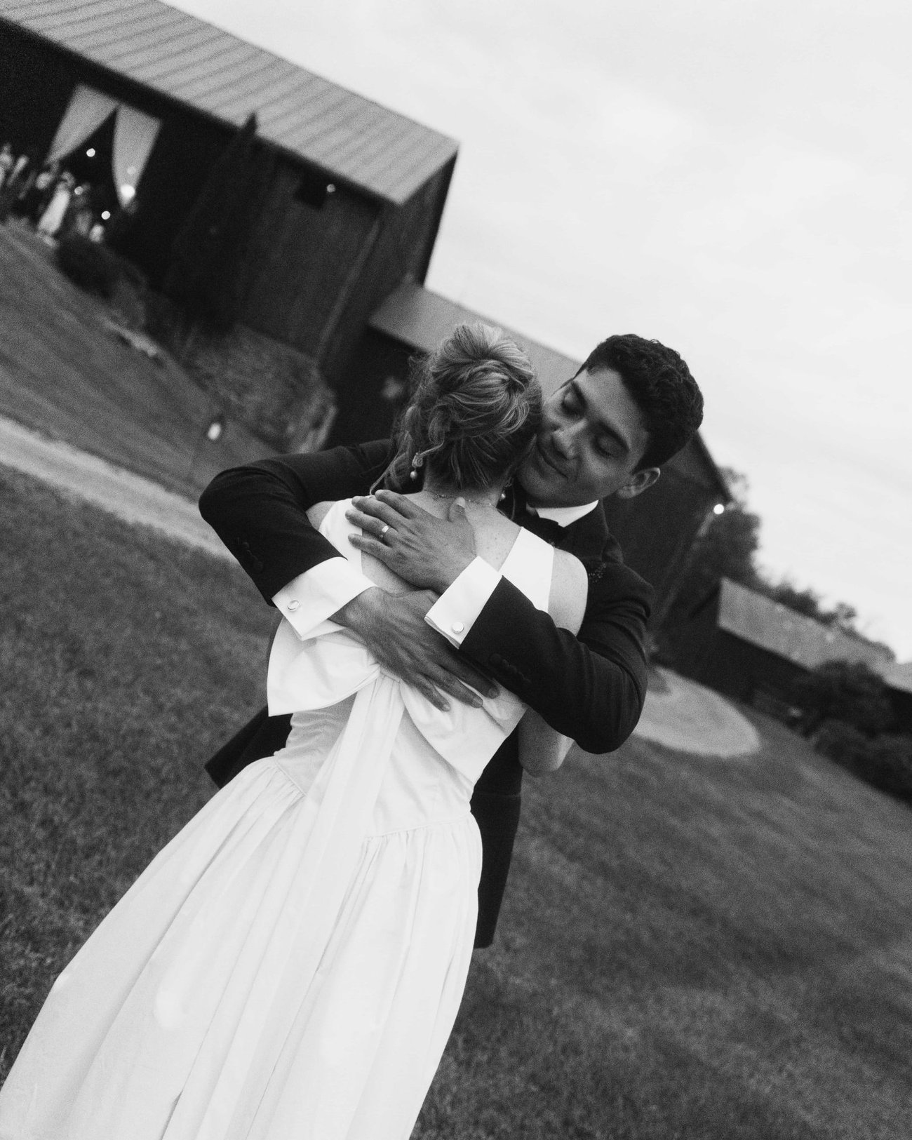 Black and white, a groom holding the bride from behind on the lawn outside a lit barn, his eyes closed against her shoulder