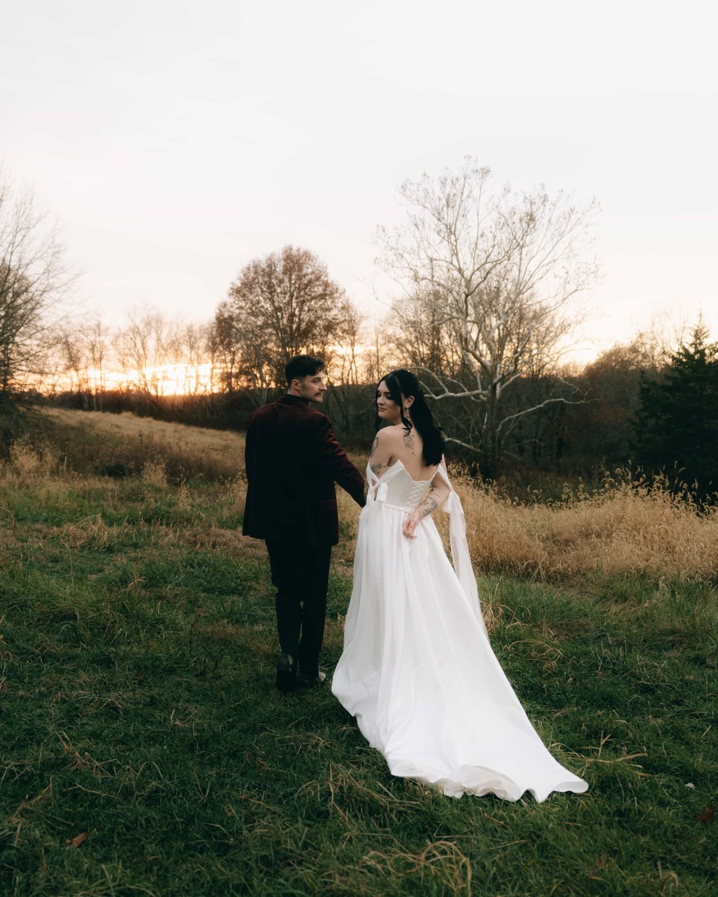 A couple walking hand in hand through a winter field at dusk, bare trees along the far edge