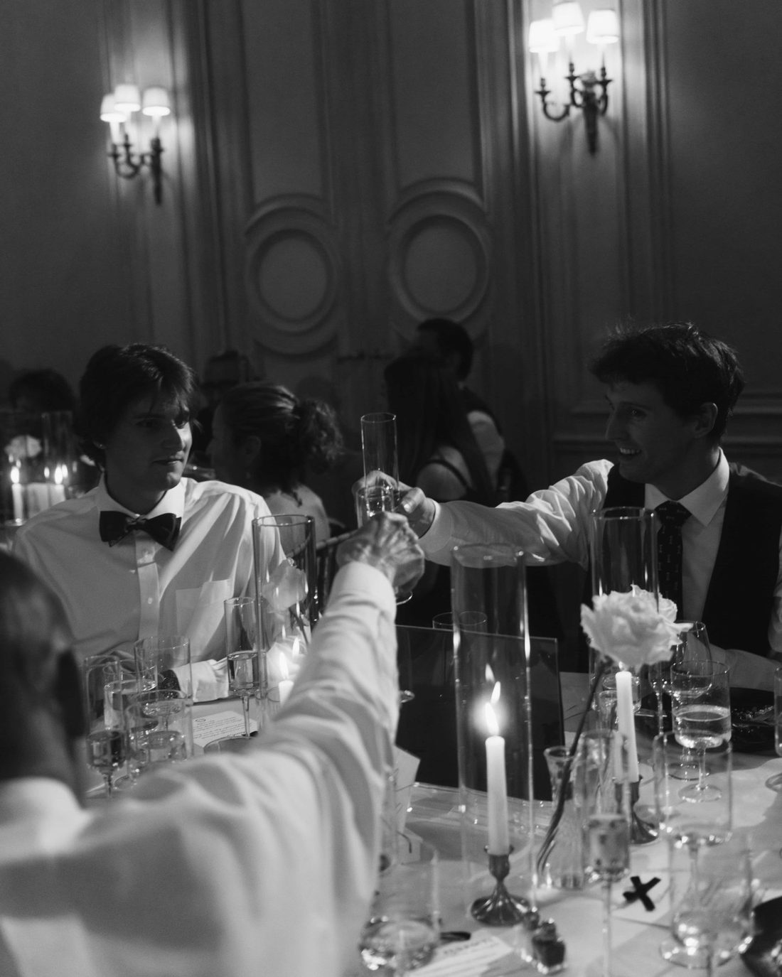 Black and white, guests reaching across a candlelit table to touch glasses in a panelled ballroom