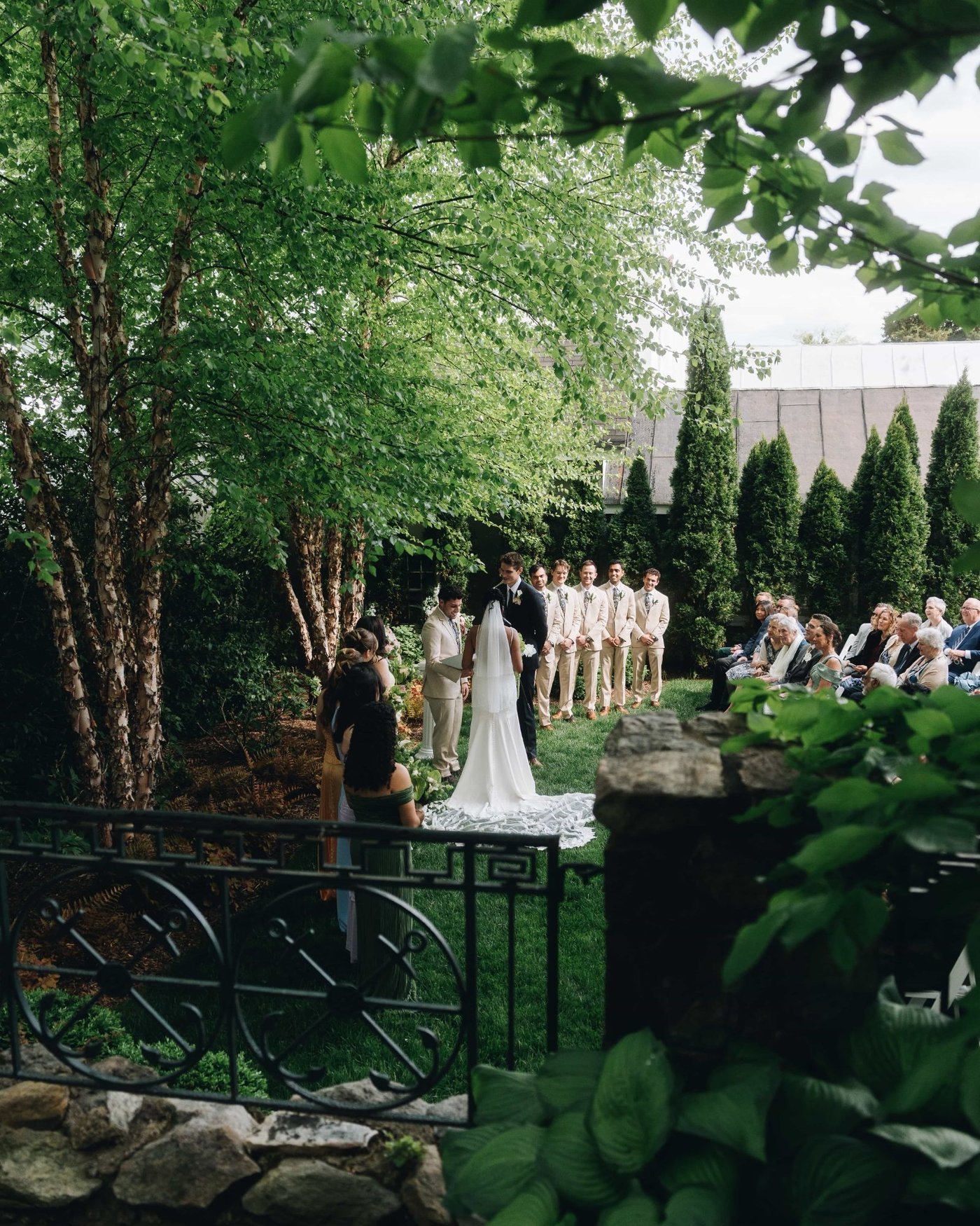A garden ceremony seen from beyond an iron railing and birch leaves, the couple at the front and the groomsmen in cream suits