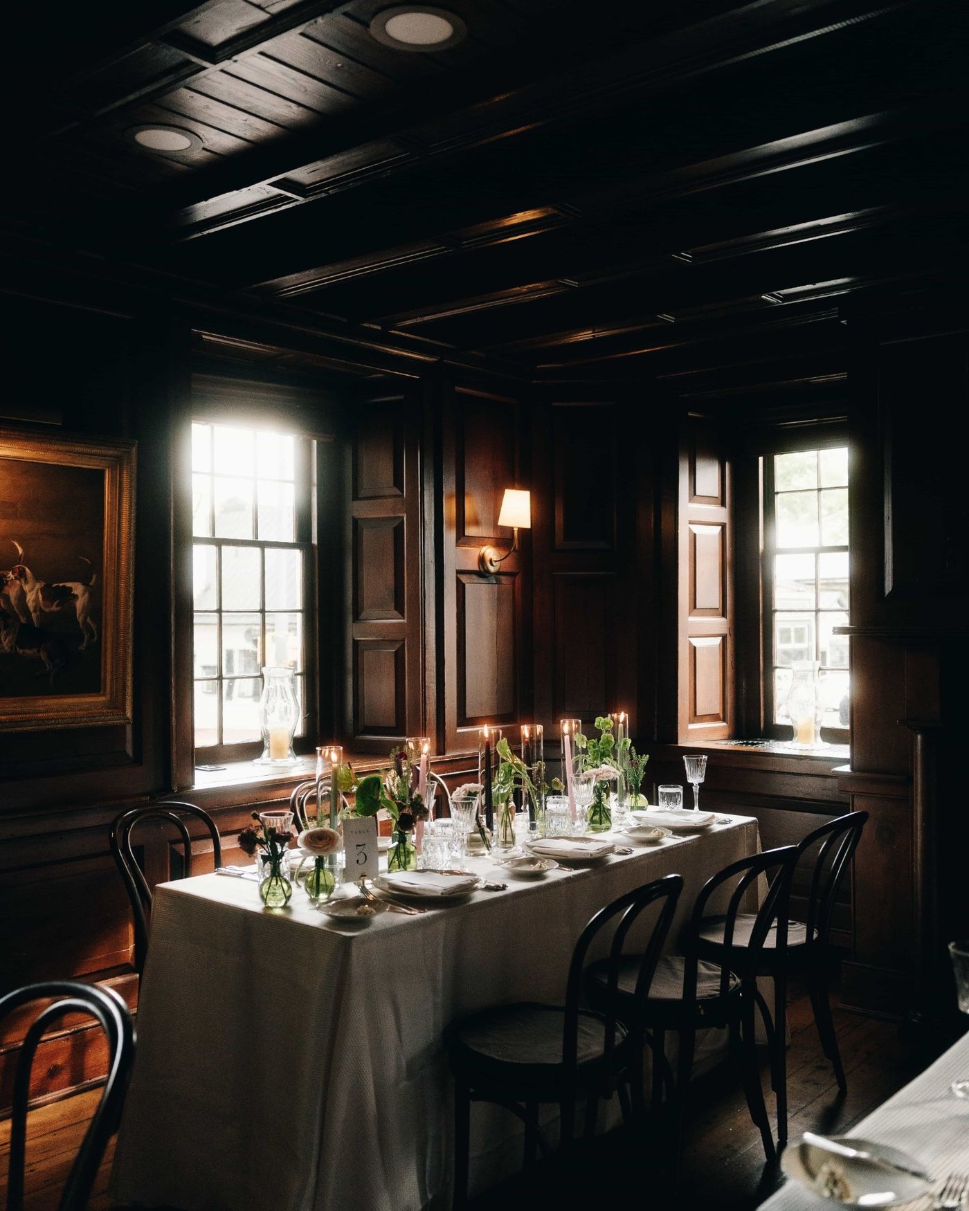 A long table laid for dinner in a dark panelled room, candles and greenery down the middle, bentwood chairs pulled in