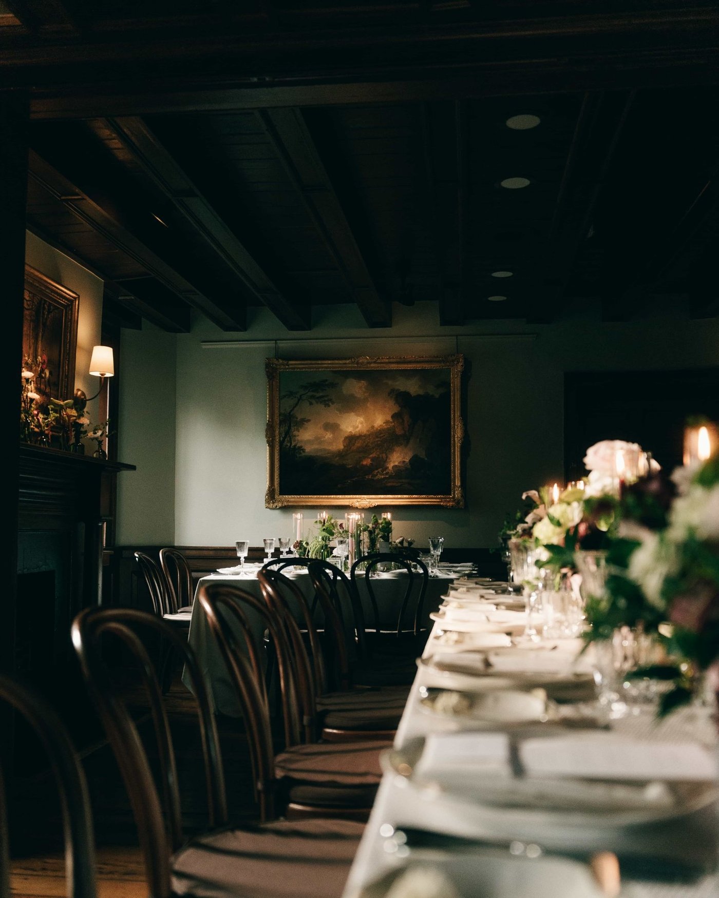 Another long table under a beamed ceiling, dressed with pale flowers, an oil painting hung above the fireplace