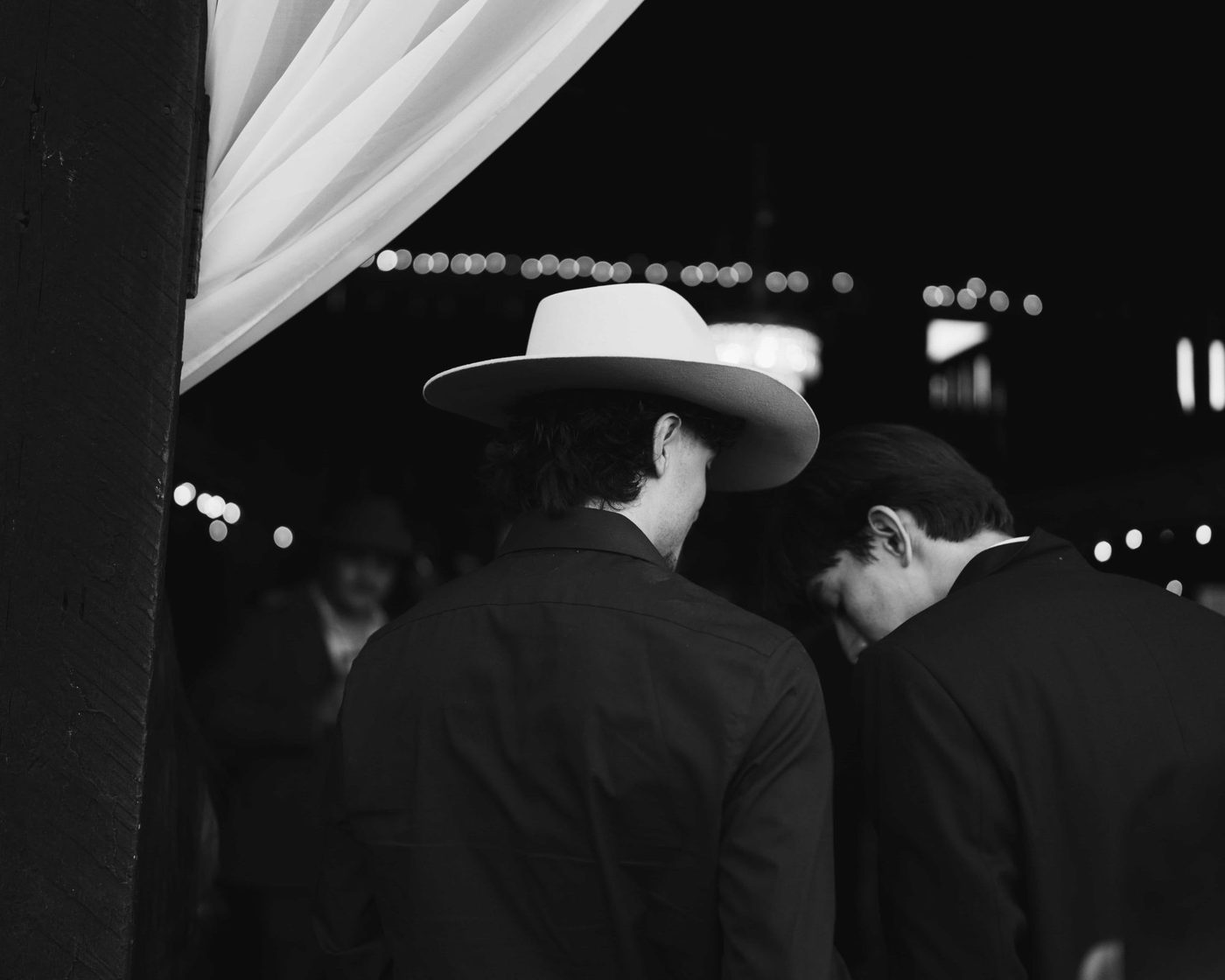 Black and white, two guests under string lights at night, one in a pale wide brimmed hat, heads bent together in conversation
