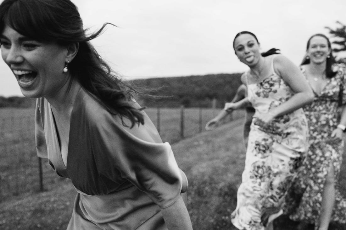 Black and white, a woman laughing as she runs across a field with two more guests running behind her