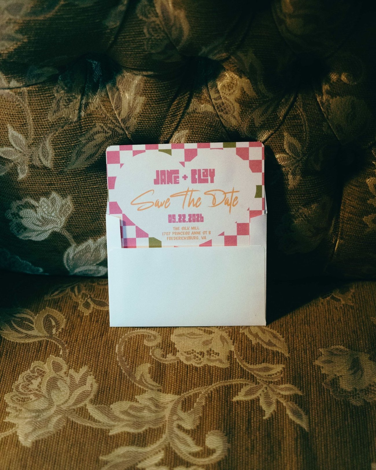 A checkerboard save the date card propped up on a tufted olive floral armchair