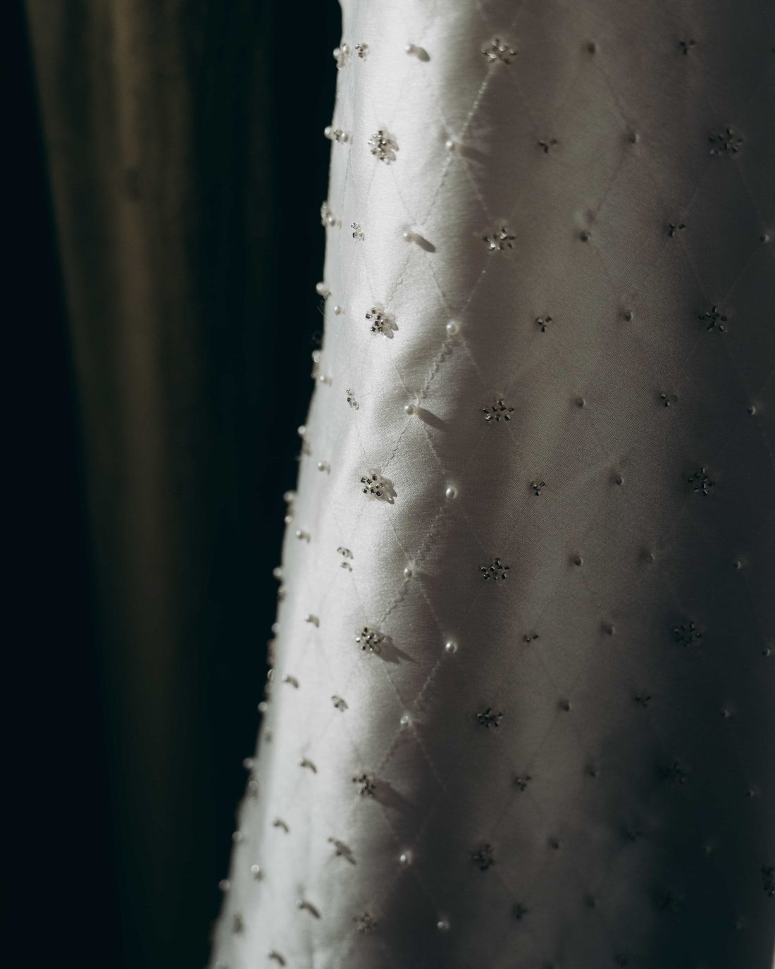 Close on a hanging gown, pearls and small crystal clusters scattered across quilted silk in a shaft of light