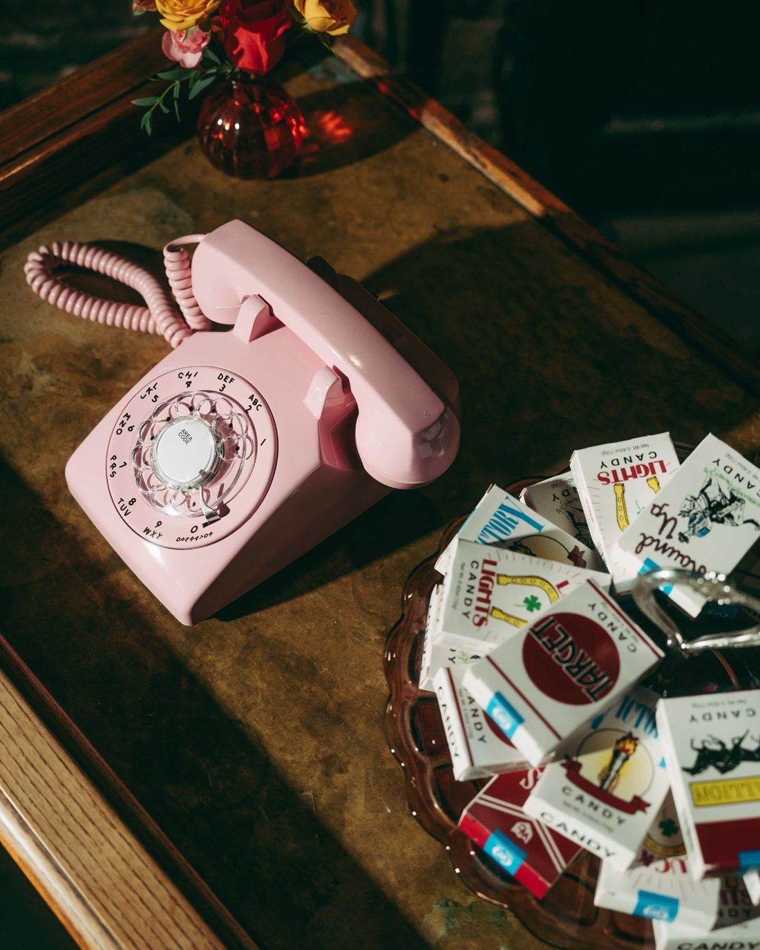 A pink rotary telephone on a worn table beside a dish heaped with candy cigarette packs