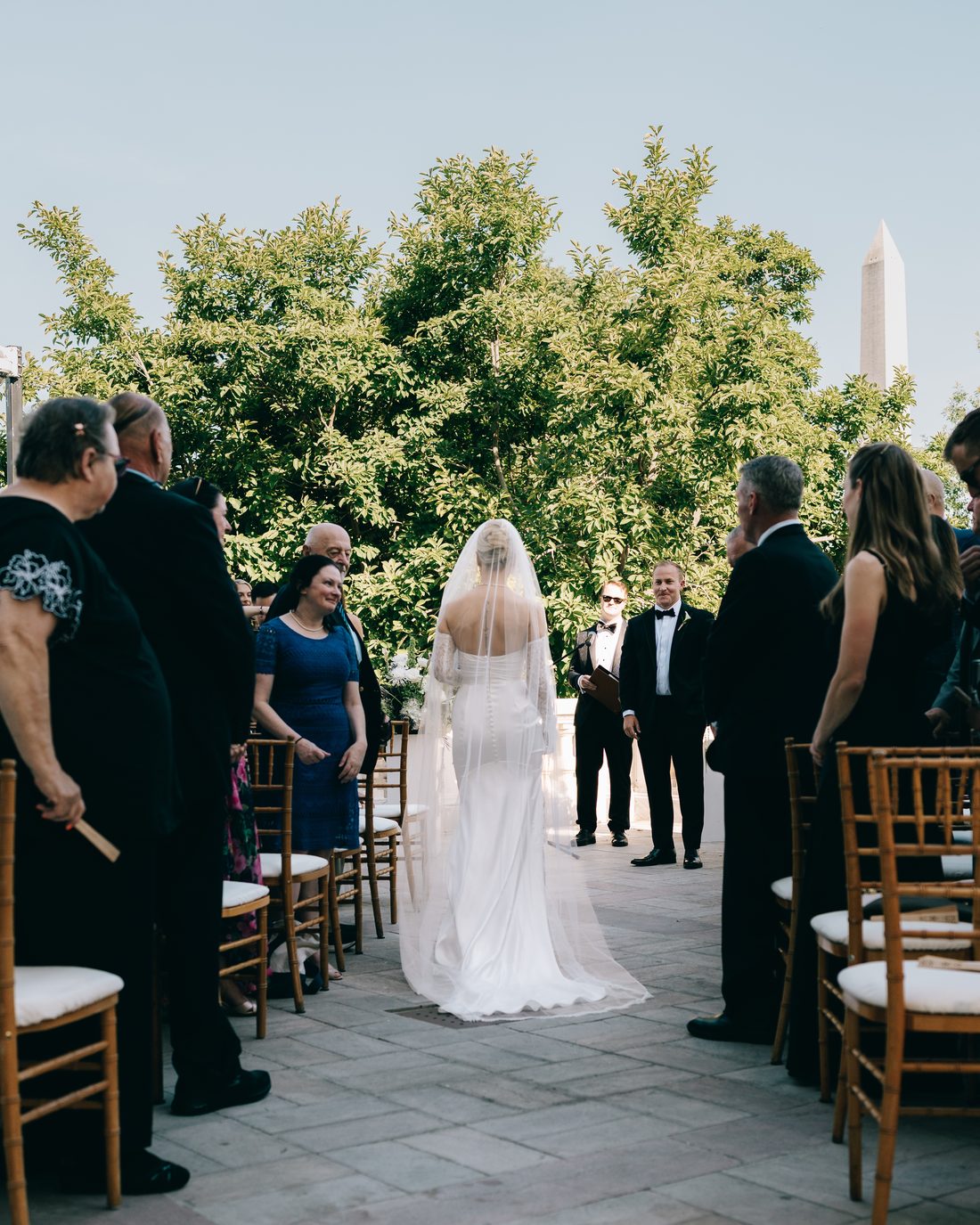 A bride seen from behind at the top of the aisle, guests on their feet and the Washington Monument rising beyond the trees