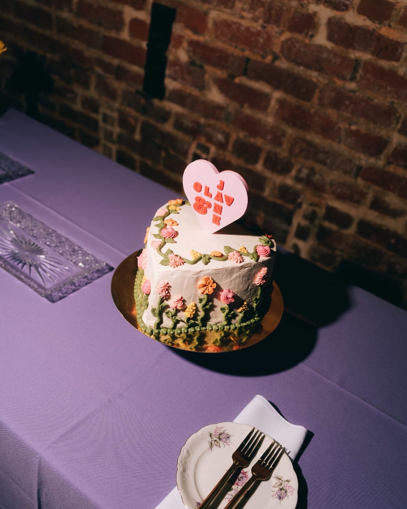 A single tier cake iced with piped flowers under a heart shaped topper, on a violet cloth against exposed brick
