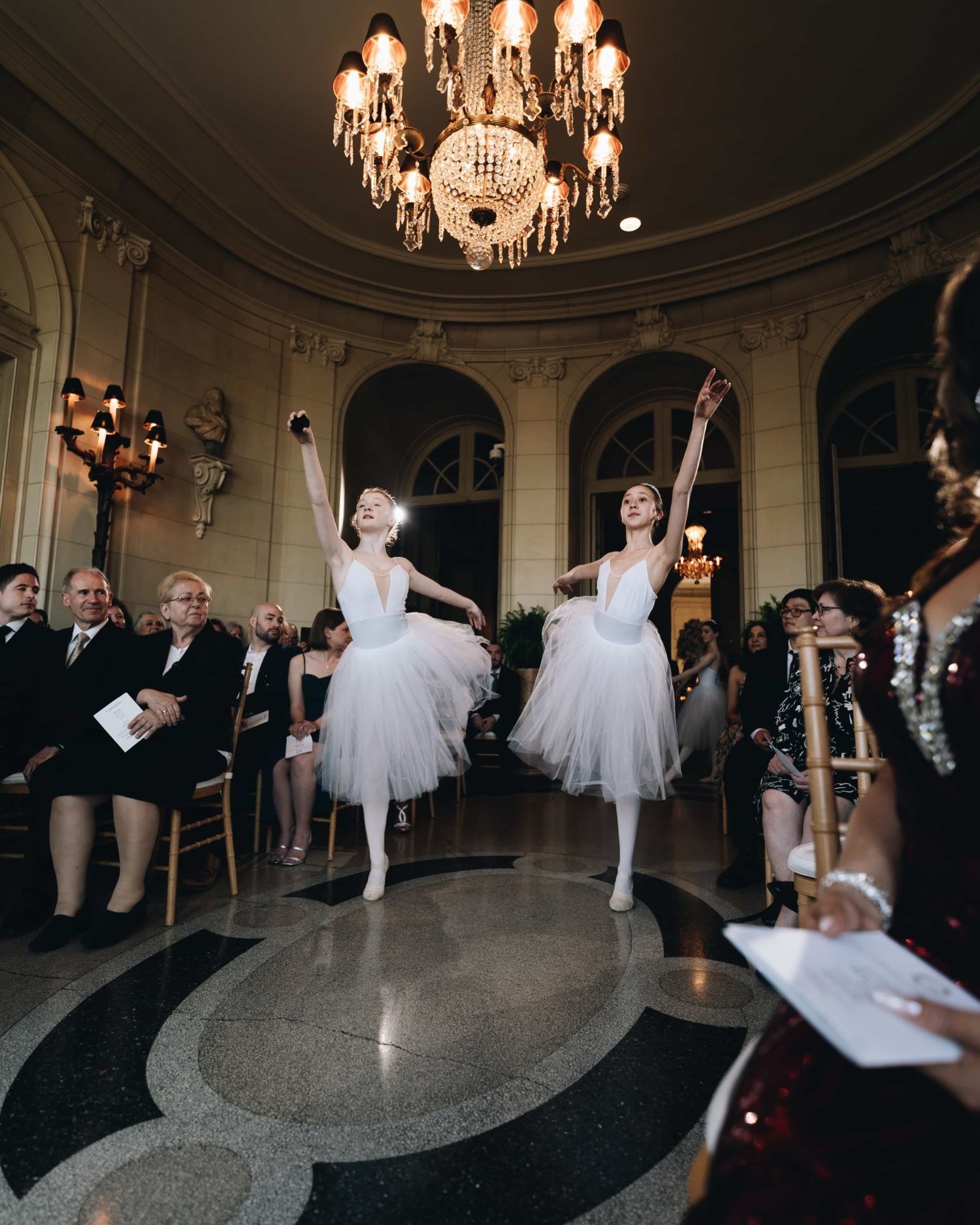 Two ballerinas dancing on a terrazzo floor beneath a chandelier while the guests watch from their seats
