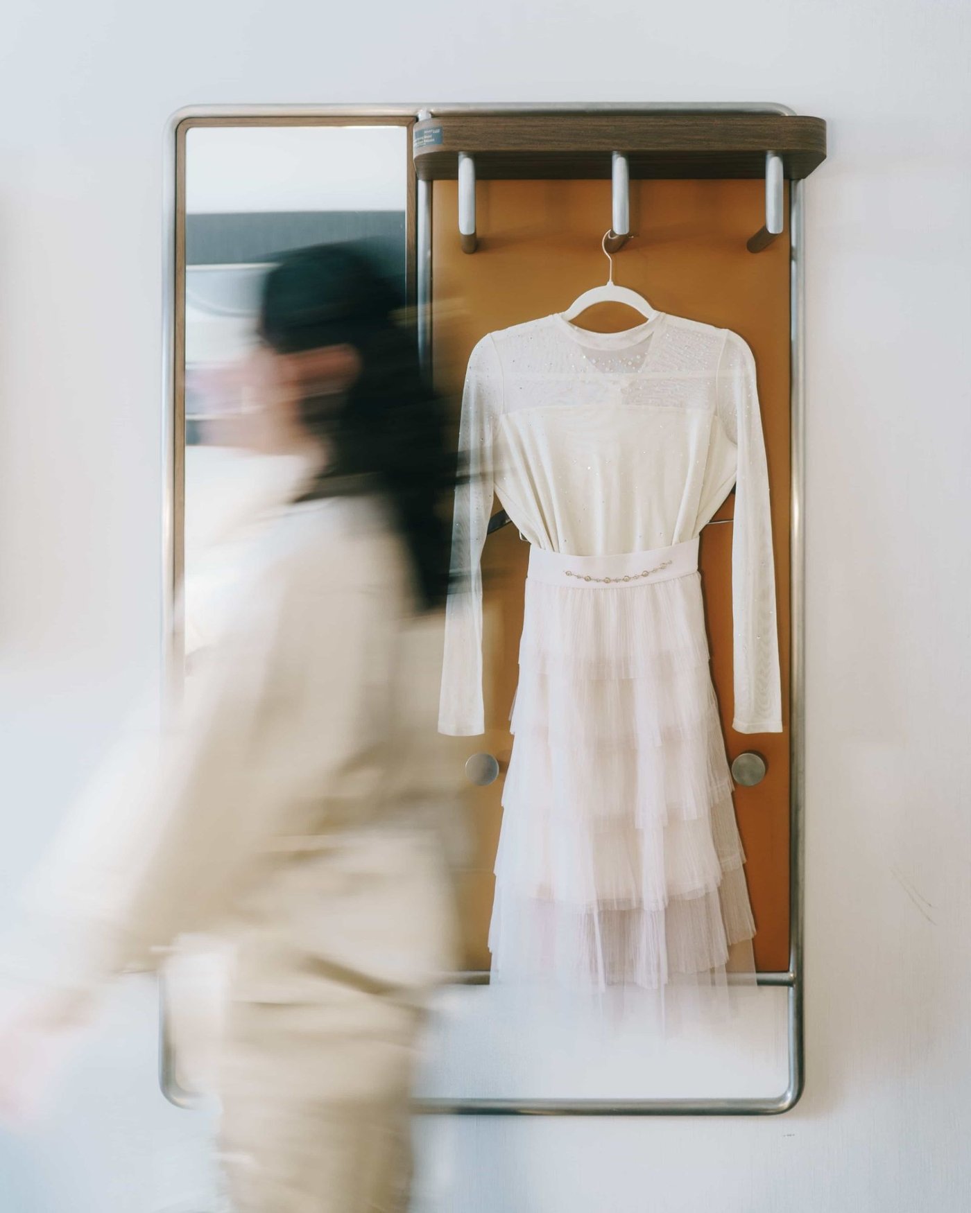 A tiered tulle dress hanging on a hook beside a mirror, someone passing in a blur of cream