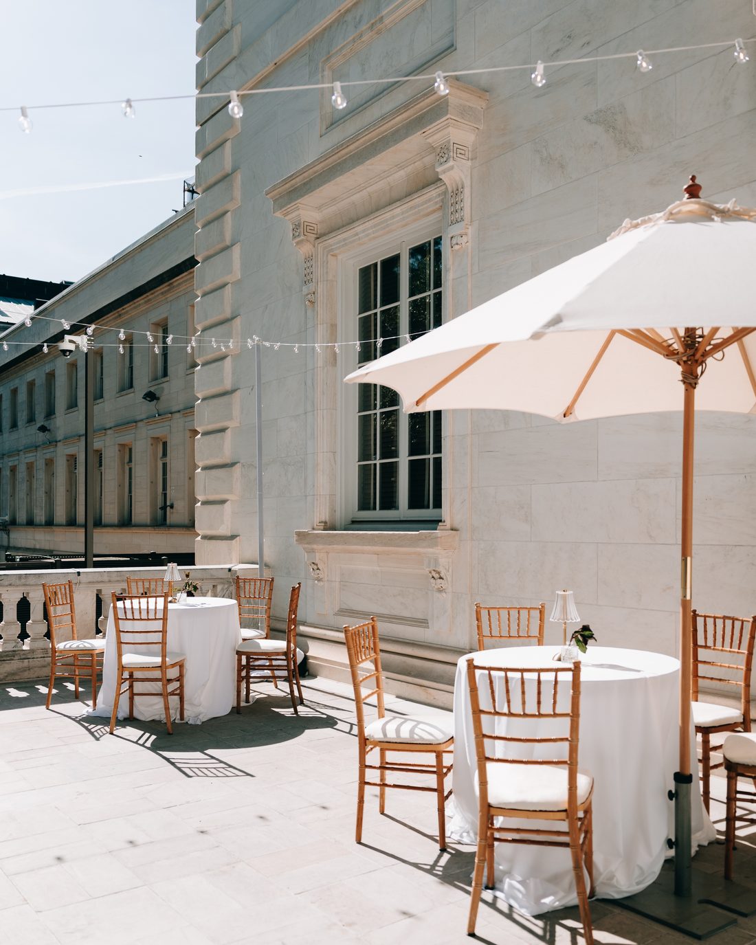 The terrace before anyone arrives: cocktail tables, gilt chairs and a canvas umbrella under strung lights